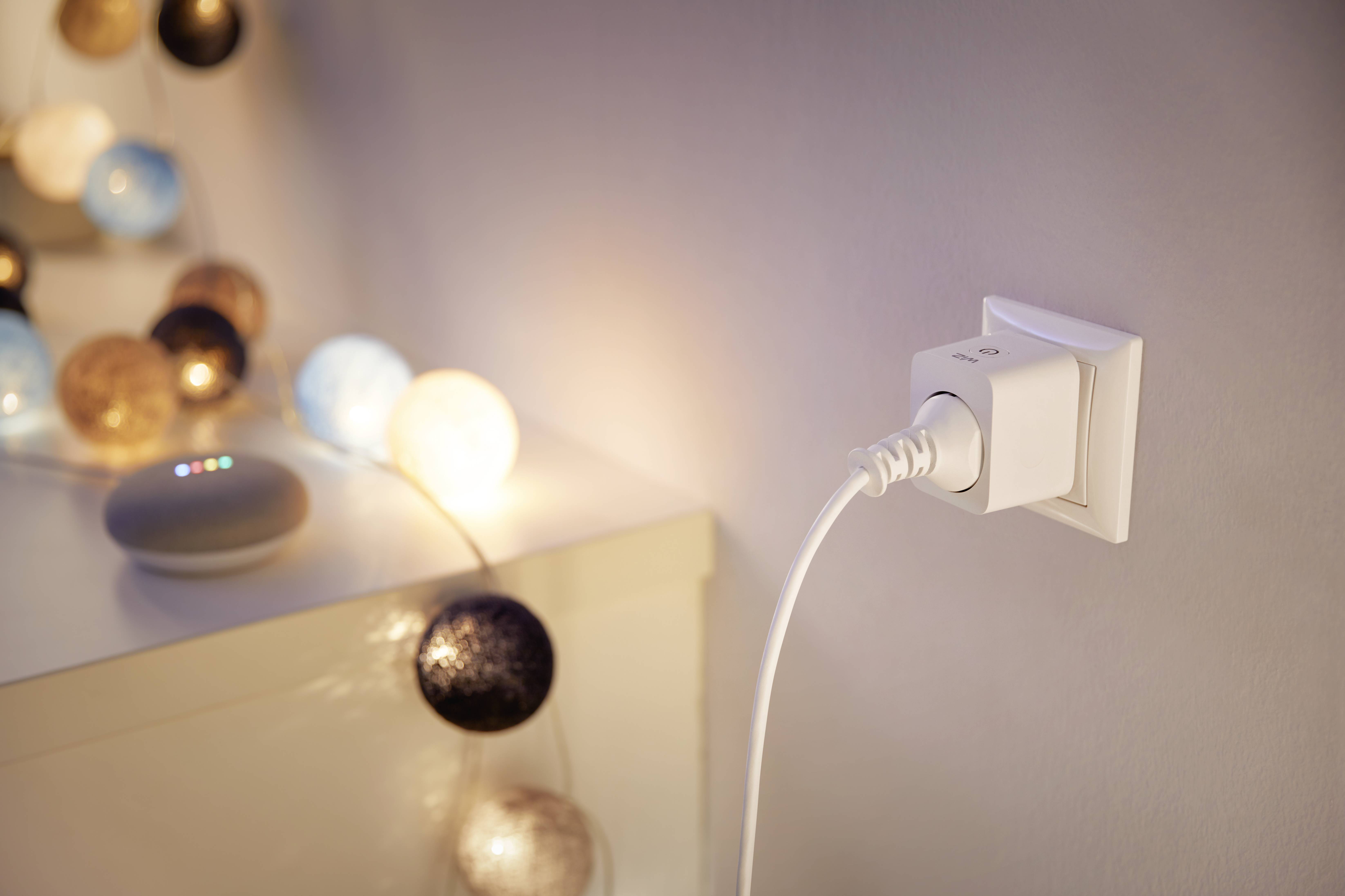 Stopcontact met ingestoken kabel, naast een slingerlamp die oplicht op een meubelstuk. Daarop rust een slim geluidsapparaat.