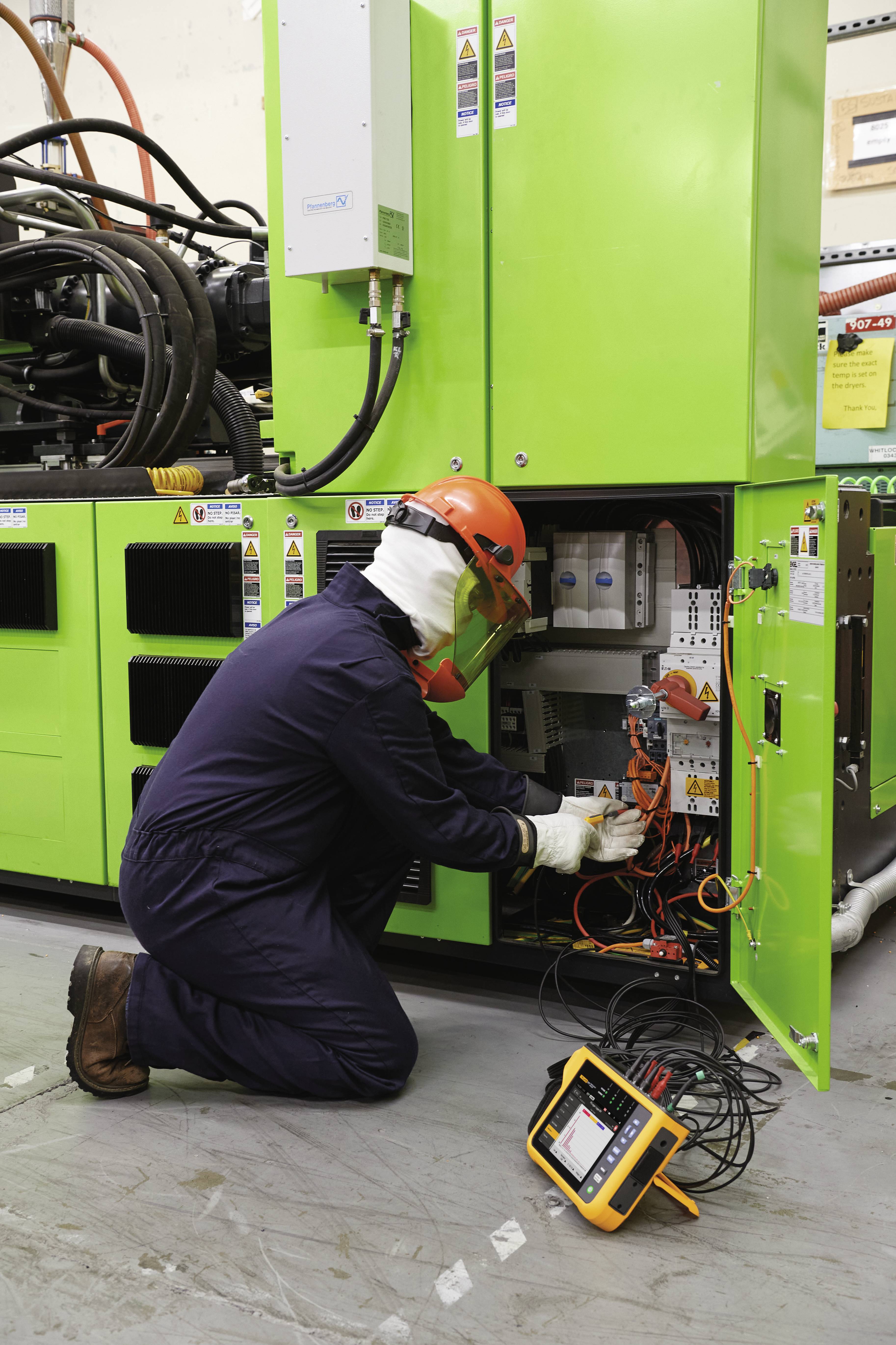 Technicus in beschermende kleding controleert machinebediening met meetapparatuur. Groene schakelkast geopend, focus op elektronische componenten.