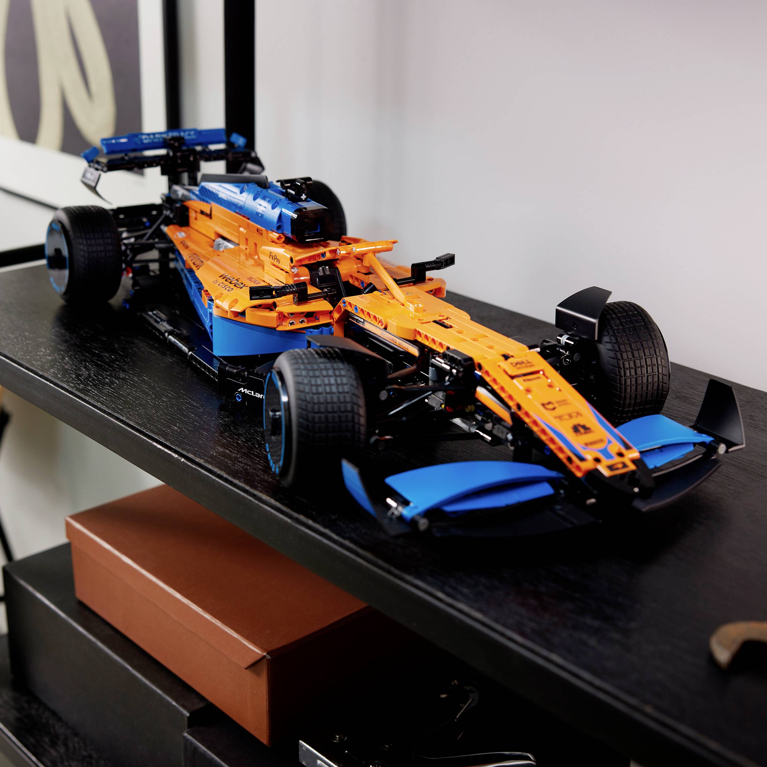 Een oranje-blauw modelauto met racewagen-ontwerp staat op een plank boven bruine dozen. Het ziet er gedetailleerd en technisch uit.