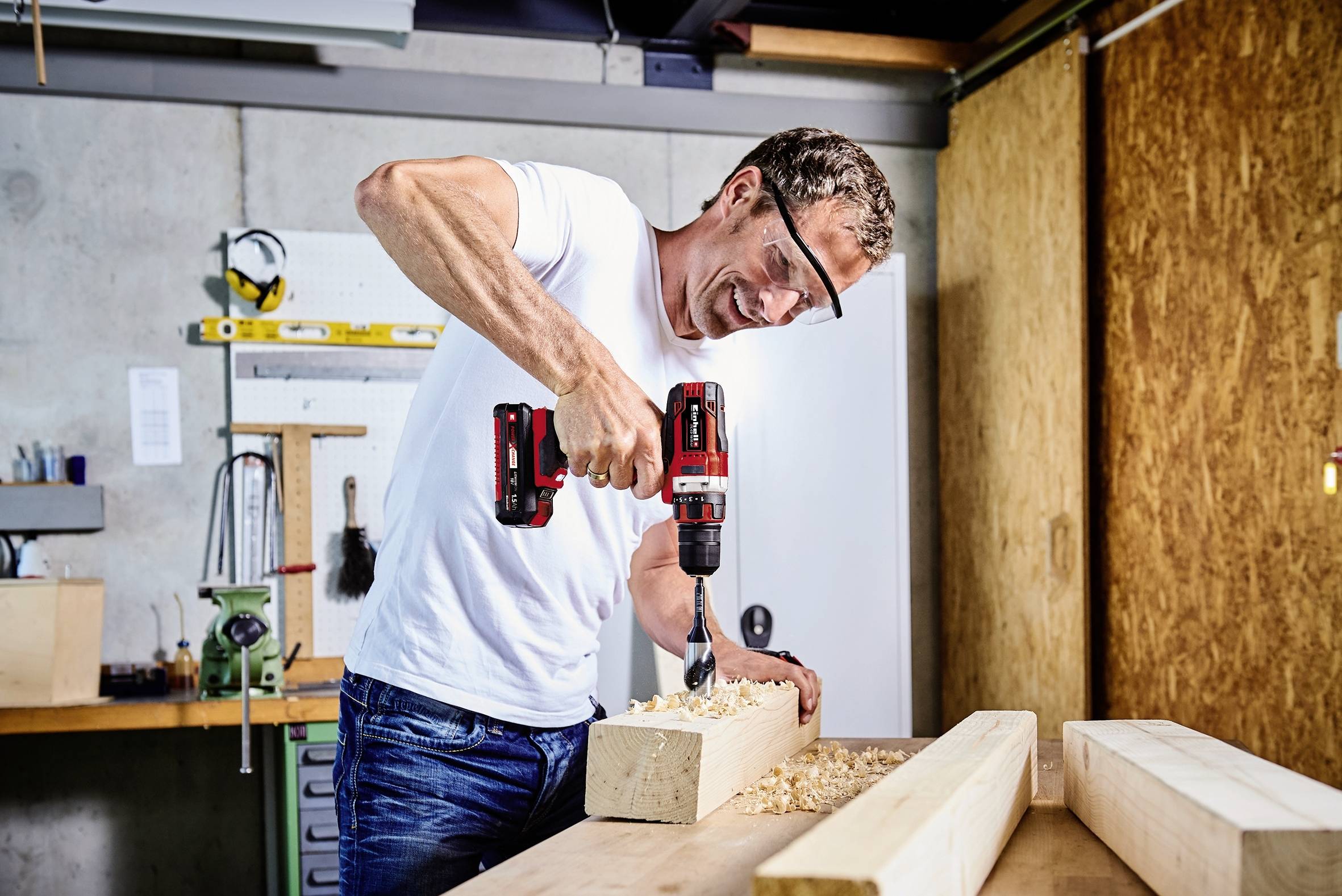 Een man lacht terwijl hij in een werkplaats een gat boort in een houten balk. Gereedschappen en houtbewerkingen zijn op de achtergrond zichtbaar.