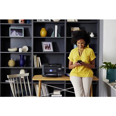 Vrouw in een gele blouse zit lachend met een mobiele telefoon in een modern kantoor voor een rek met decoratie en een printer op tafel.