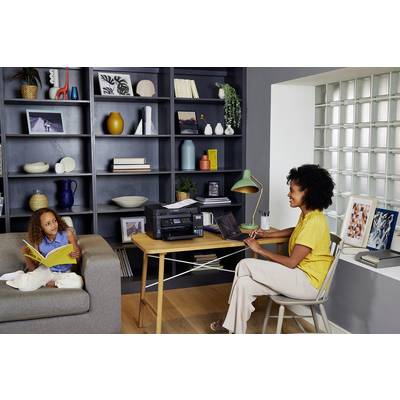 Een vrouw werkt op een laptop aan een bureau met een printer, terwijl een kind op de bank zit te lezen. In de achtergrond staat een boekenrek.