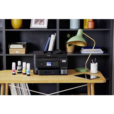 Op een houten tafel staat een printer met inktflesjes ernaast. Op de achtergrond zijn een boekenkast en een groene lamp zichtbaar.
