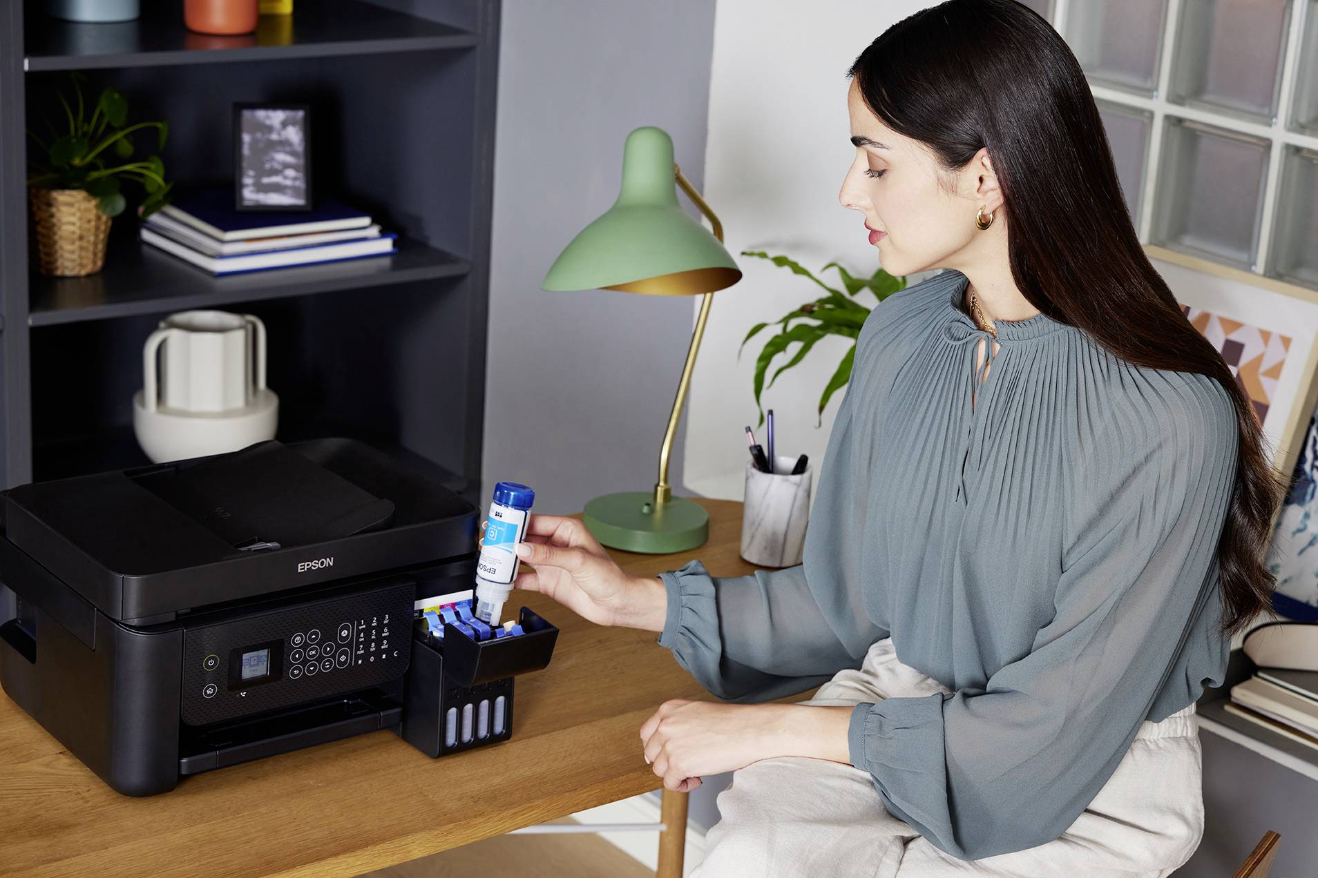 Een vrouw vult inkt in een printercartridge. De printer staat op een bureau, op de achtergrond zijn planten en lampen.