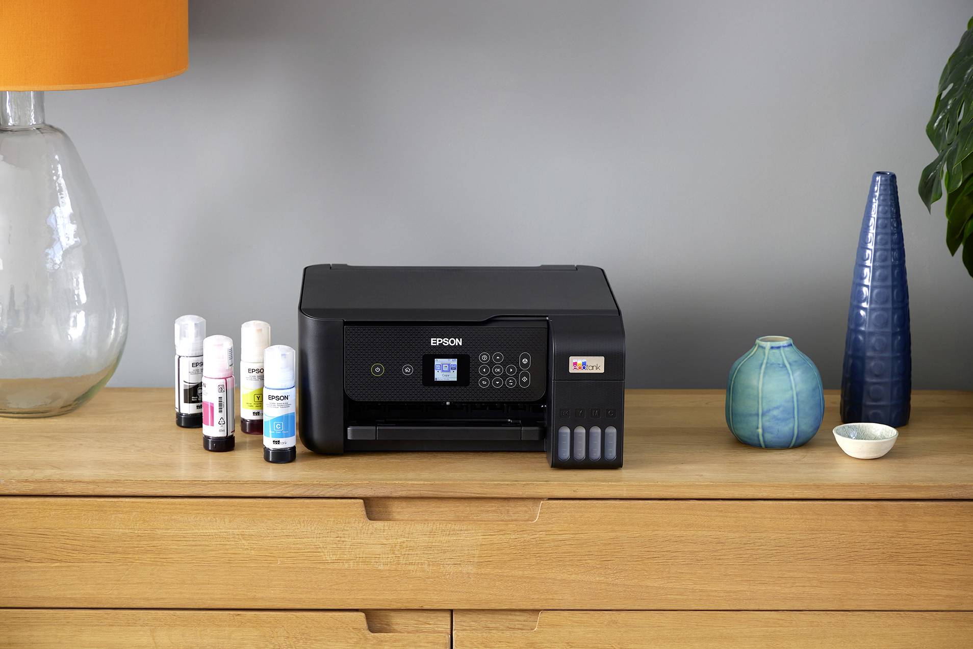 Een zwarte Epson-printer staat op een lichtbruine houten tafel, omringd door inktflesjes en decoratieve vazen.