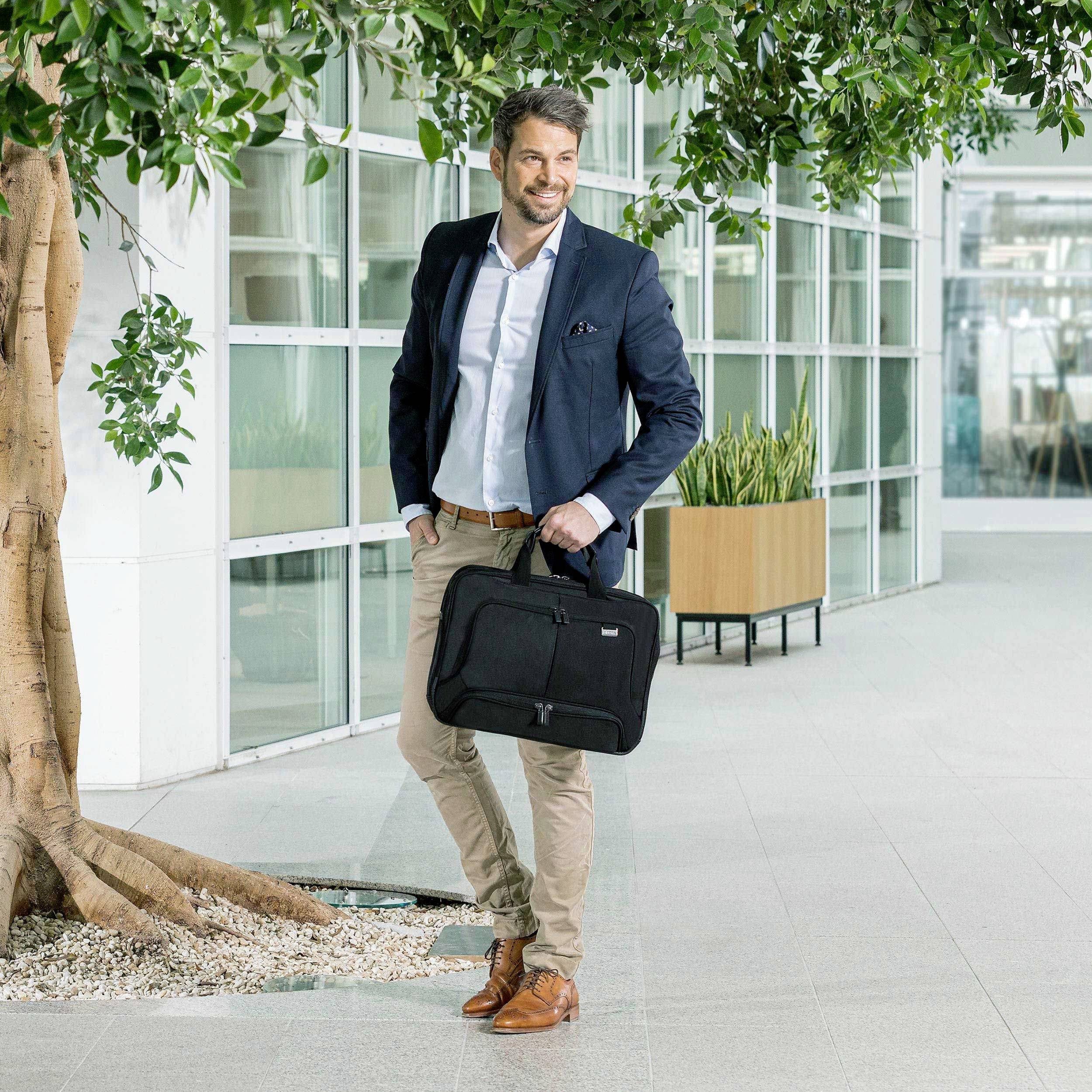 Een man in zakelijke kleding loopt glimlachend door een modern kantoorgebouw. Hij draagt een zwarte laptoptas in zijn rechterhand.