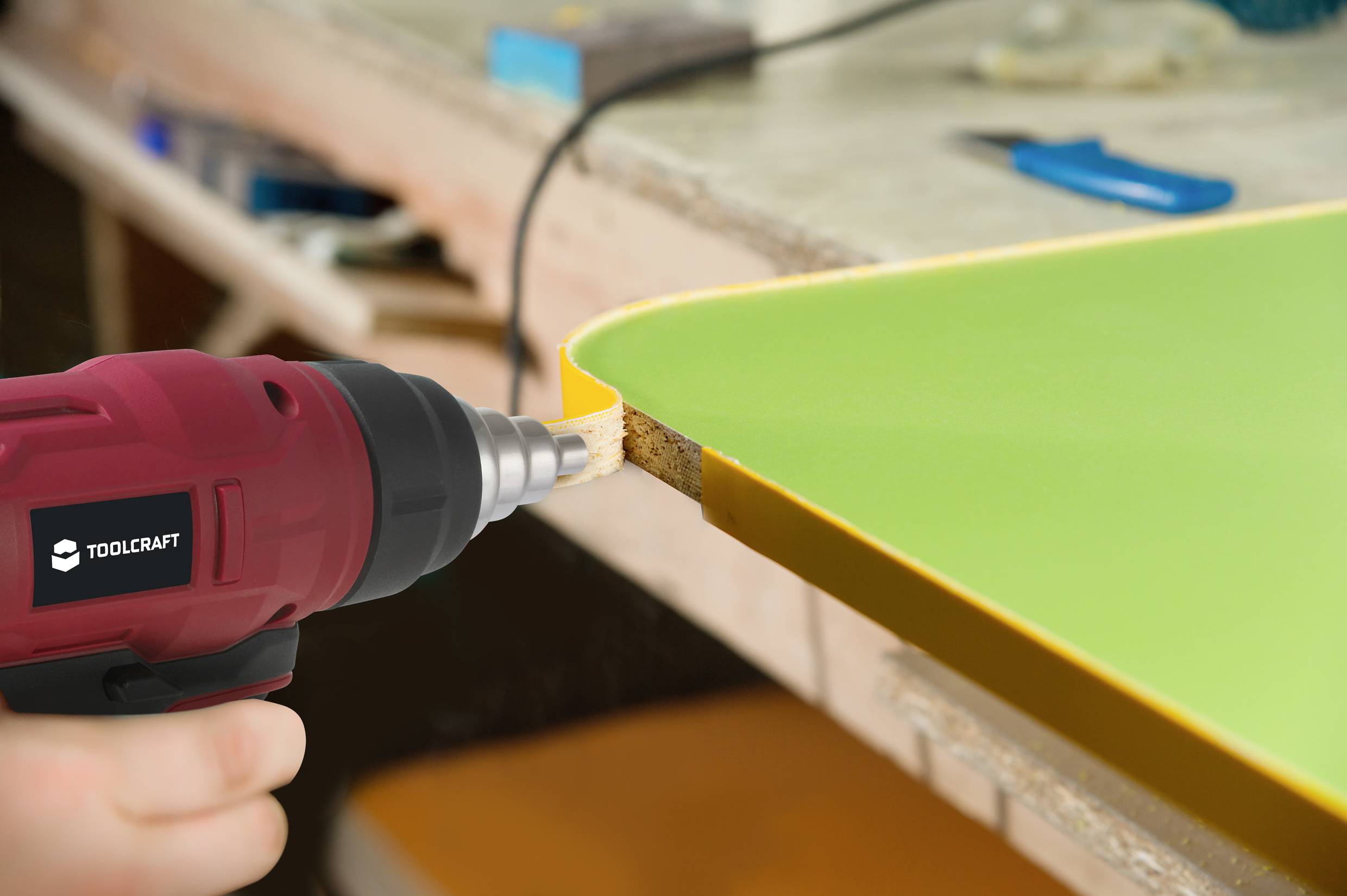 Een rode boormachine van TOOLCRAFT bewerkt de rand van een groene tafelblad. Op de achtergrond is een werkbank te zien.