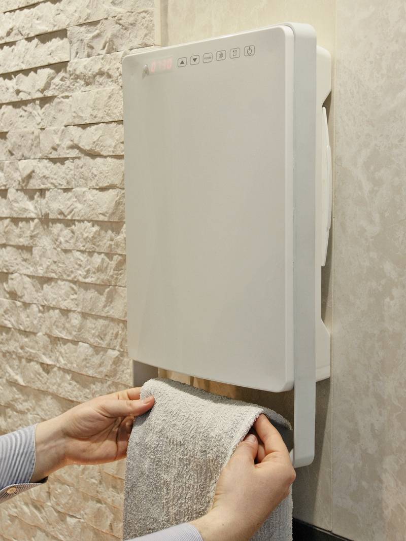Een handdroger blaast lucht op een handdoek die wordt vastgehouden door een persoon. De droger is bevestigd aan een stenen muur.