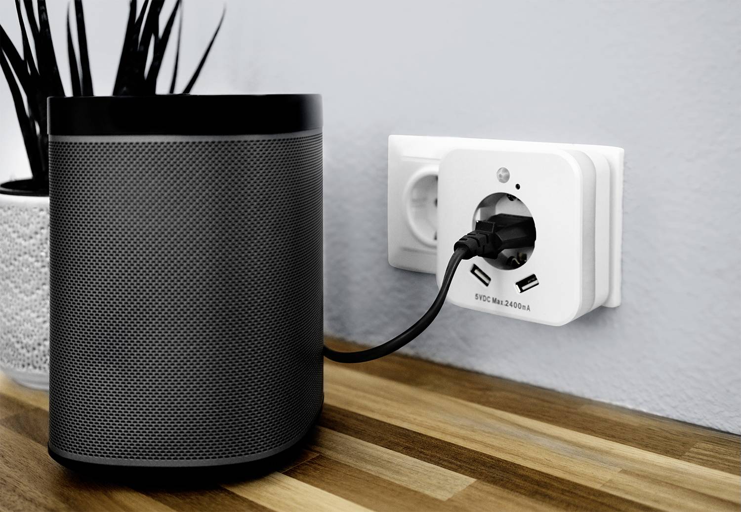 'Een intelligente speaker is aangesloten op een stopcontact dat is gemonteerd op een houten oppervlak. Op de achtergrond een plant.'