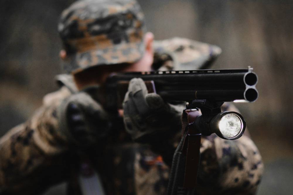 Een persoon in camouflage-uitrusting richt een jachtgeweer. De omgeving is wazig, de focus ligt op de loop van het wapen.