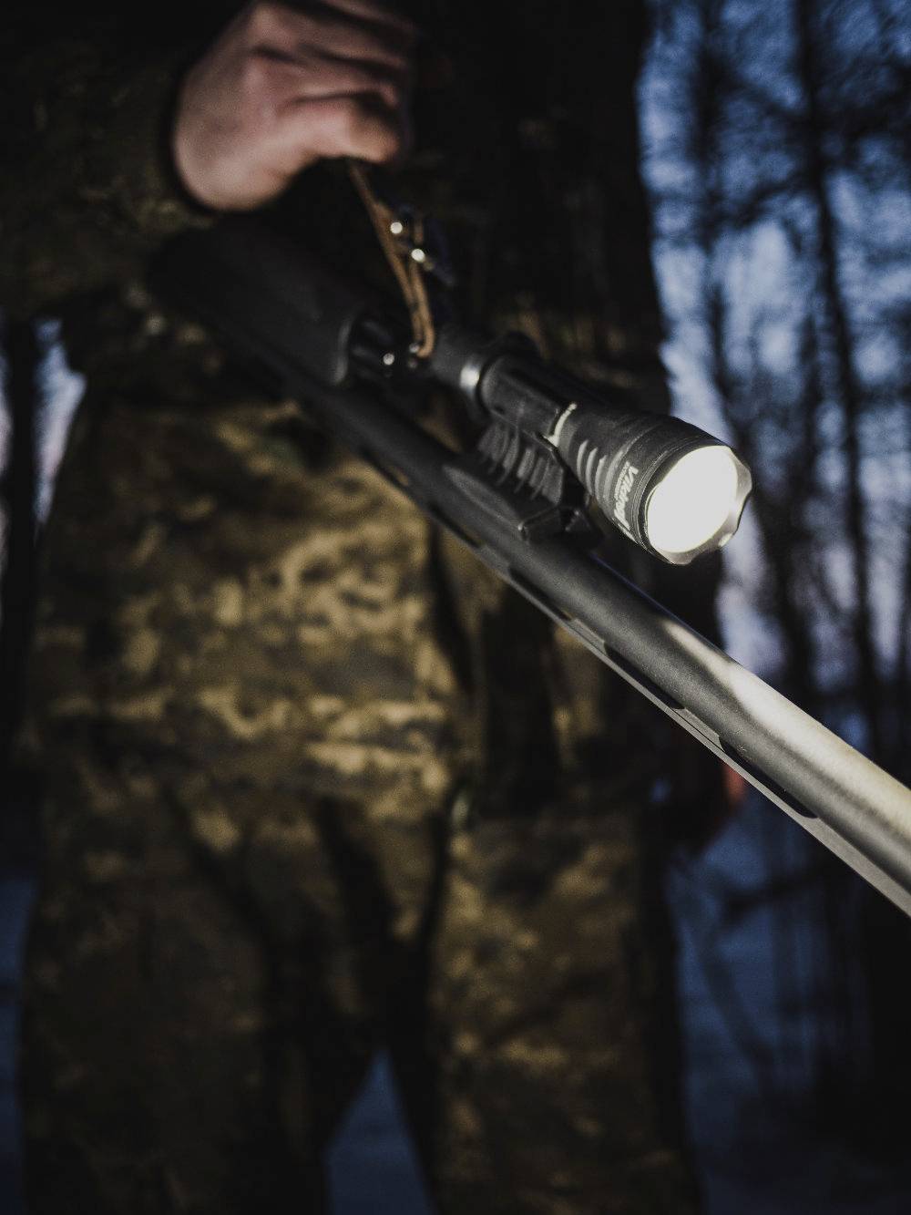 Een persoon in camouflage-uitrusting houdt een geweer vast met een gemonteerde zaklamp, met onscherpe bomen op de achtergrond bij schemering.