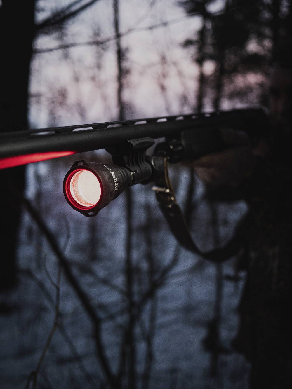 Een persoon in het bos houdt een vuurwapen vast met een rood licht ingeschakeld. Achtergrond toont wazig silhouet van bomen bij schemeringslicht.
