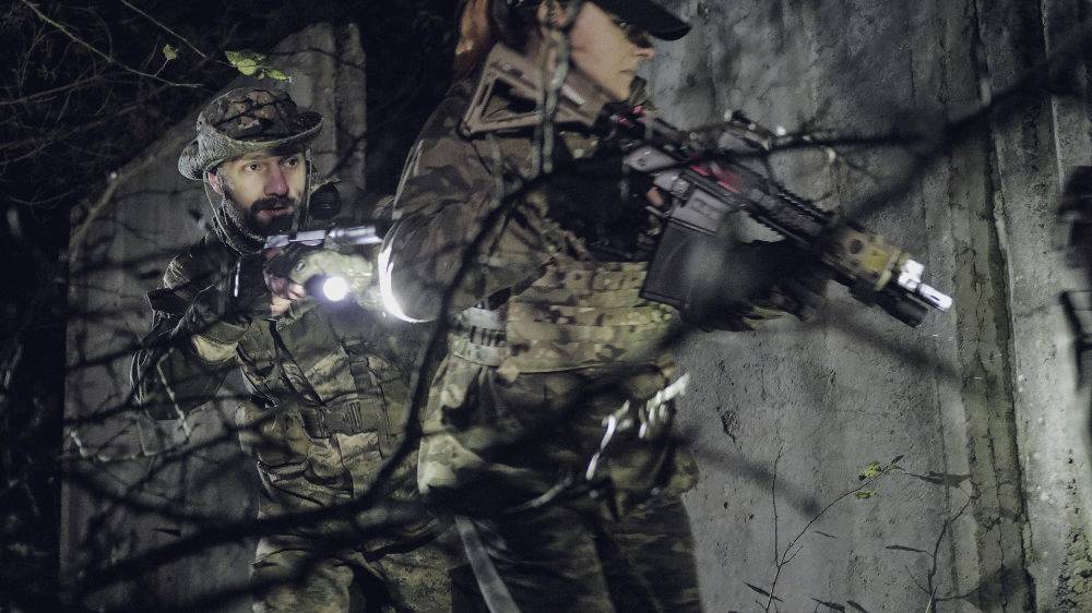 Twee personen in camouflage-uitrusting met geweren en zaklantaarns doorzoeken een donkere omgeving, als onderdeel van een tactische operatie.