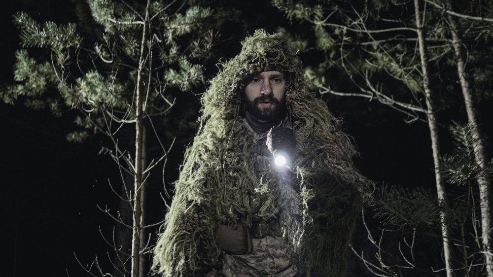 Persoon, gekleed in camouflage en met een zaklamp, staat 's nachts in een bos.