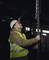 Een arbeider met veiligheidshelm en reflecterend vest inspecteert een zware ketting in een fabriekshaal bij weinig licht.
