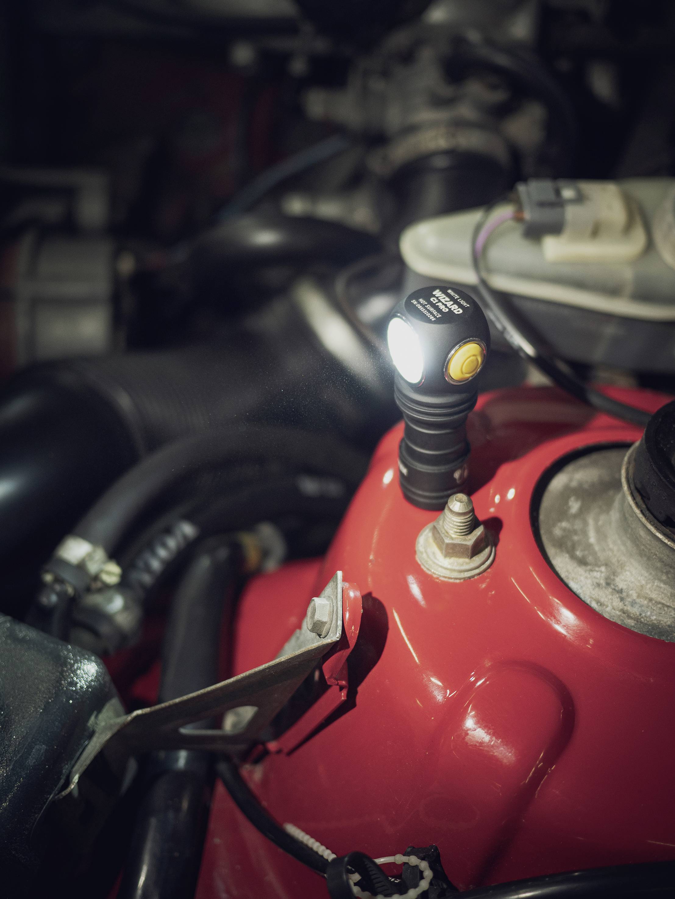 Klein LED-lampje met flexibele hals, bevestigd aan een rode metalen oppervlakte, verlicht delen van een motorruimte.