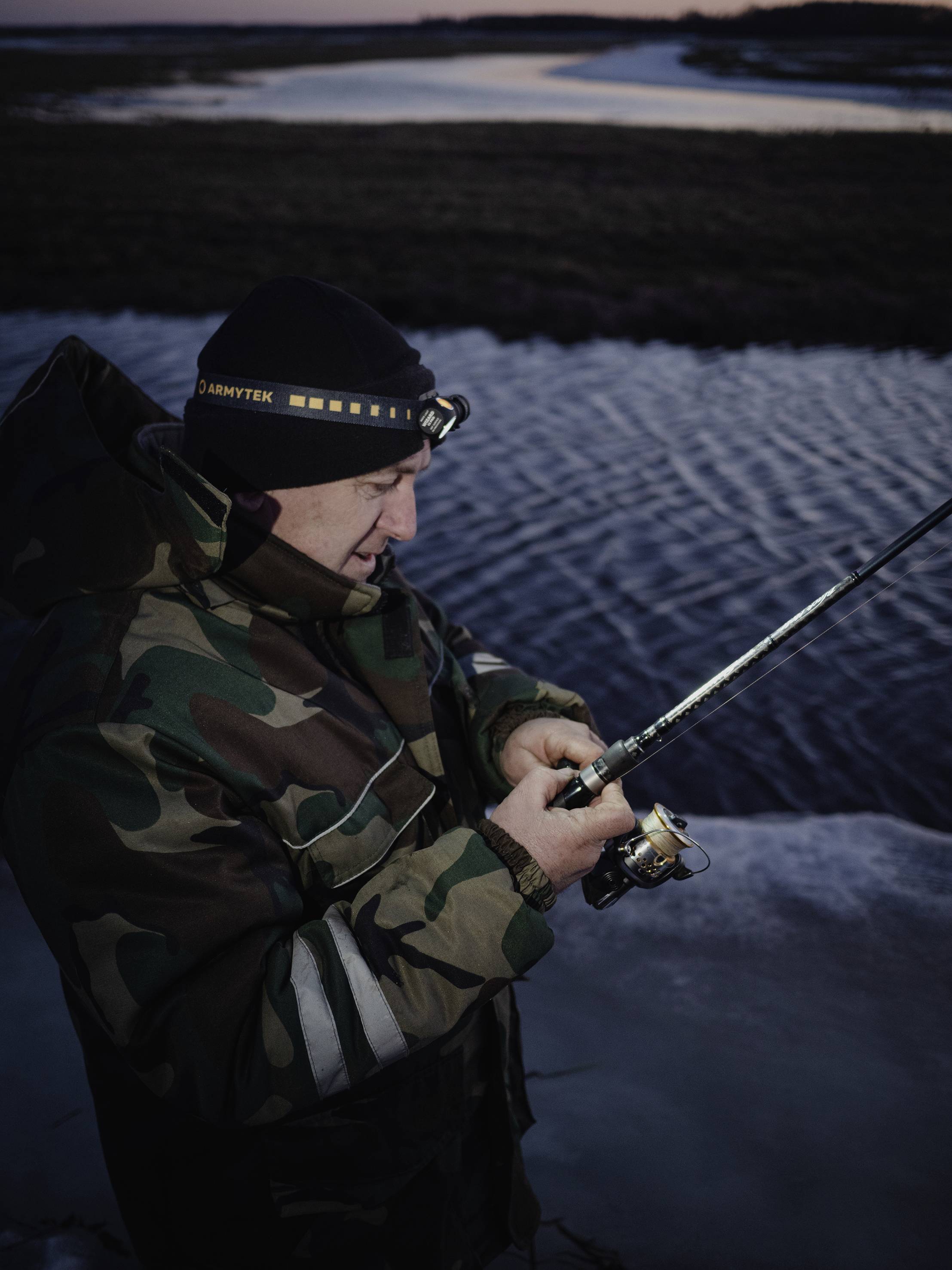 Een persoon in camouflage-kleding vist bij schemering aan een rivier. Ze draagt een hoofdlamp en concentreert zich op de hengel.