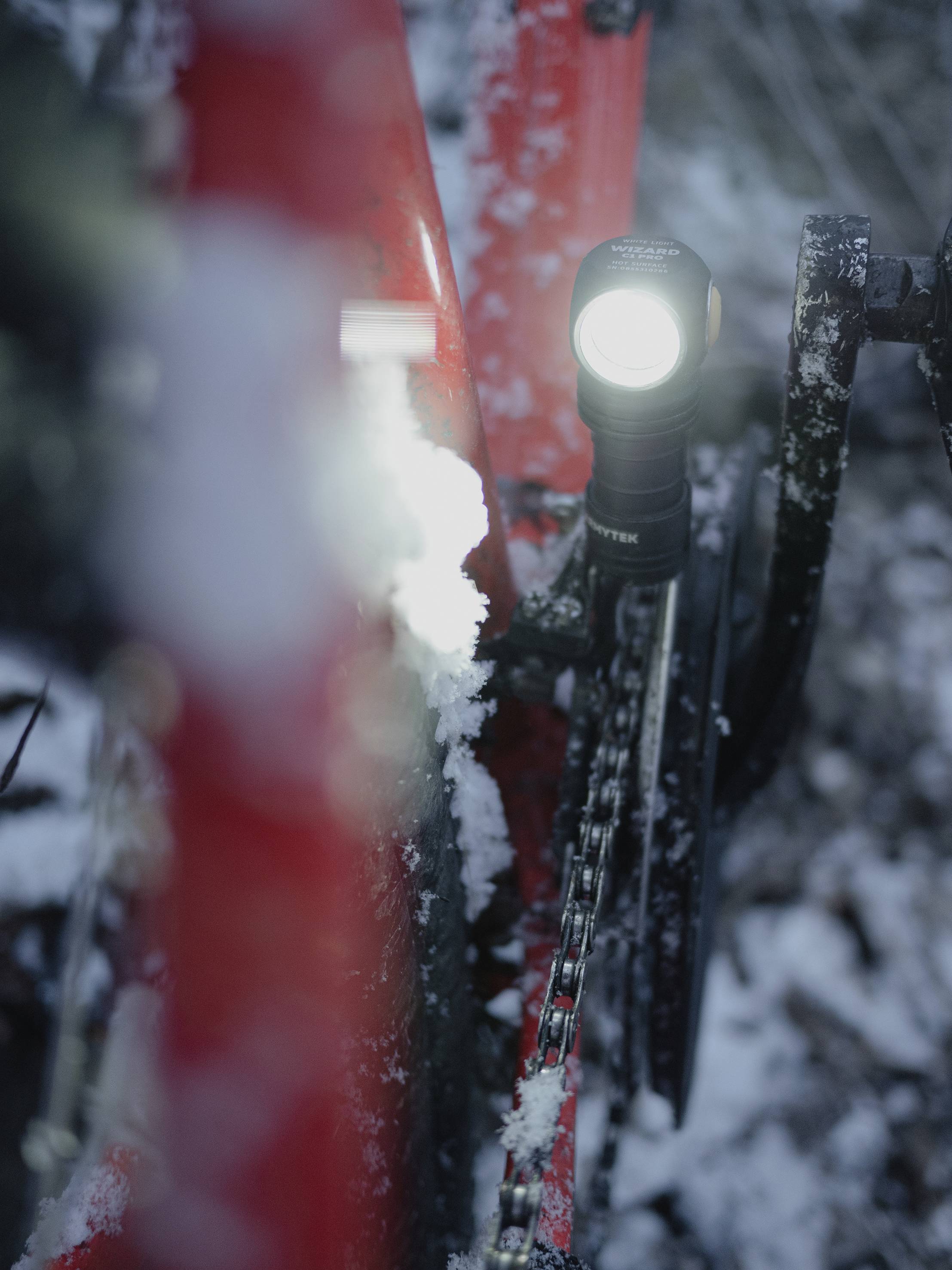 Rode fiets in de sneeuw met ingeschakerd voorlicht, zadel en ketting zichtbaar. Winterlandschap op de achtergrond.