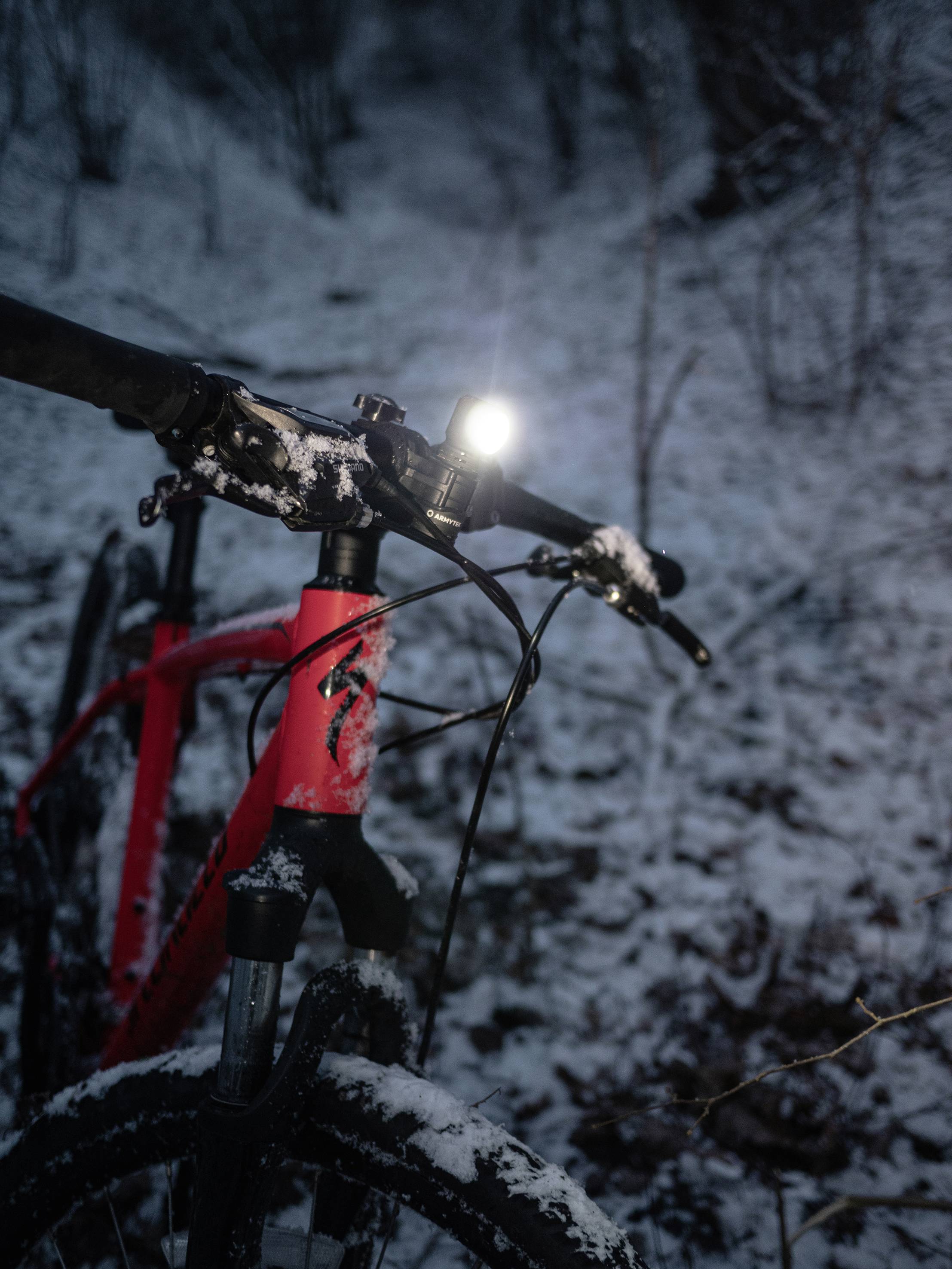 Een rood fiets met ingeschakelde voorverlichting staat in een met sneeuw bedekte bos. De omgeving is donker, alleen het licht schijnt.