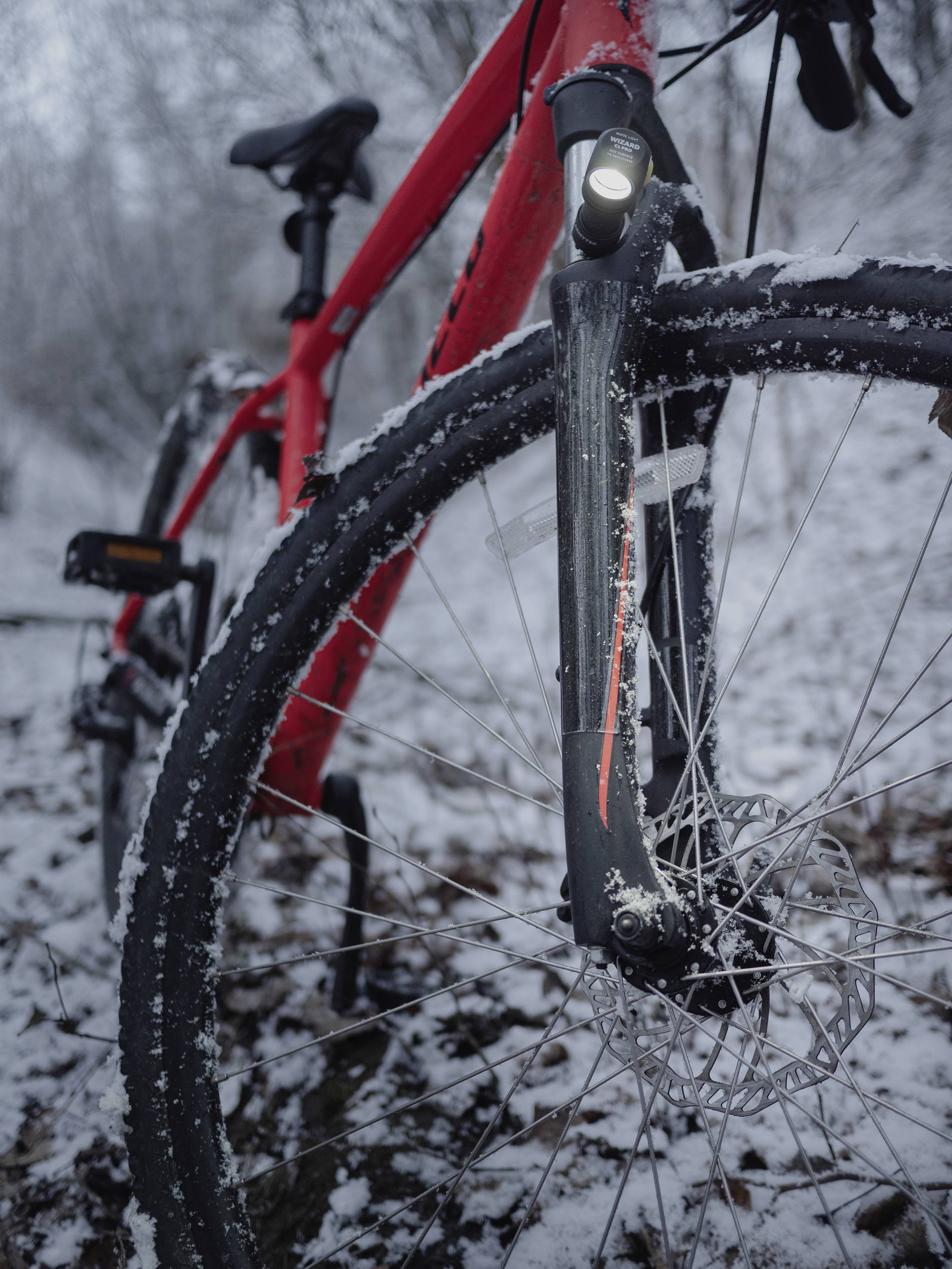 Een rode fiets met aangestoken licht staat op een met sneeuw bedekte bosweg. De close-up toont het voorwiel en sneeuw op het frame.