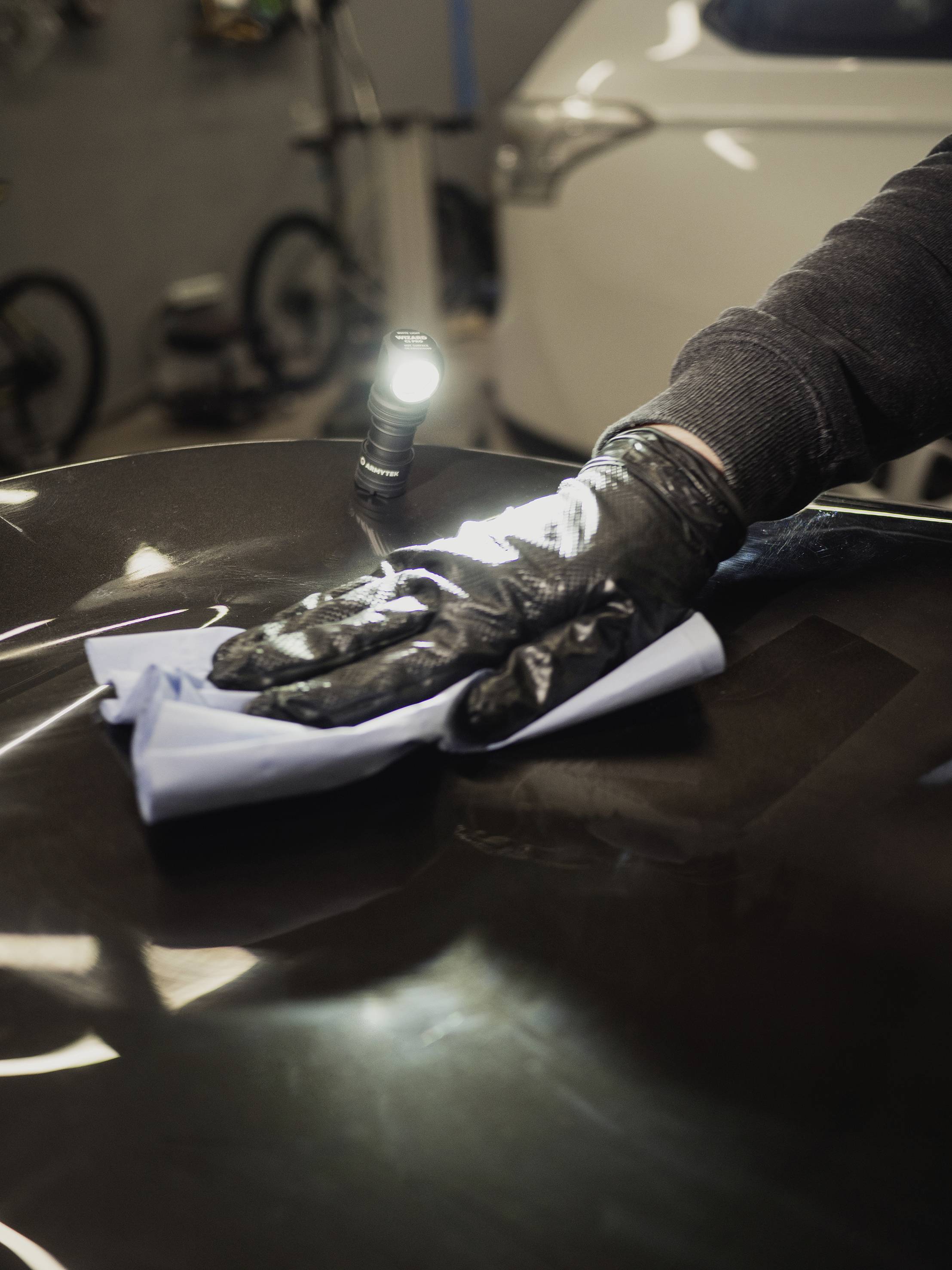 Een persoon reinigt autolak met een doek in een werkplaats; een zaklamp verlicht het oppervlak. Auto en fiets op de achtergrond.