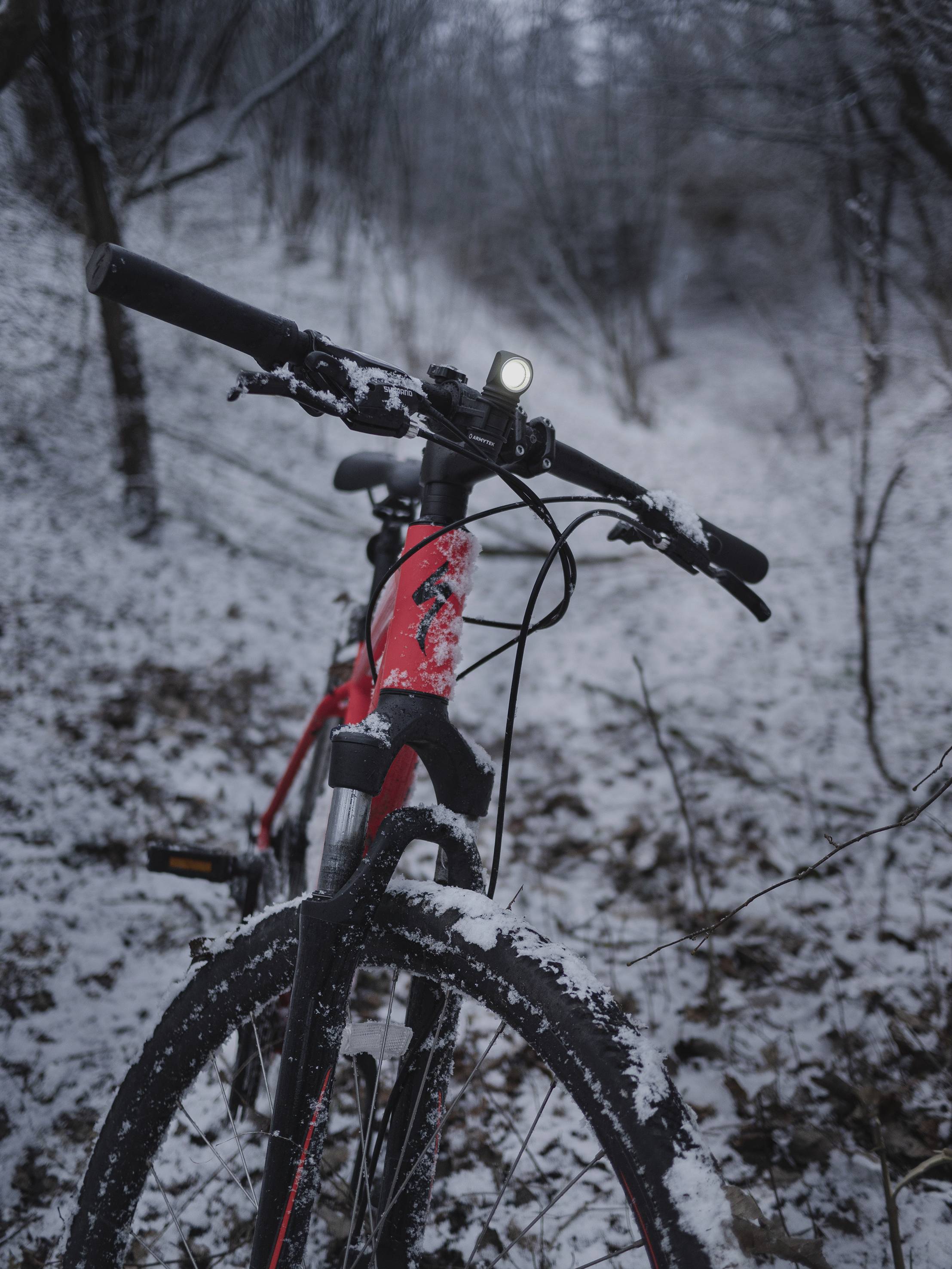 Een rode mountainbike staat op een besneeuwd bospad. Het is winter, en de fietsverlichting schijnt.