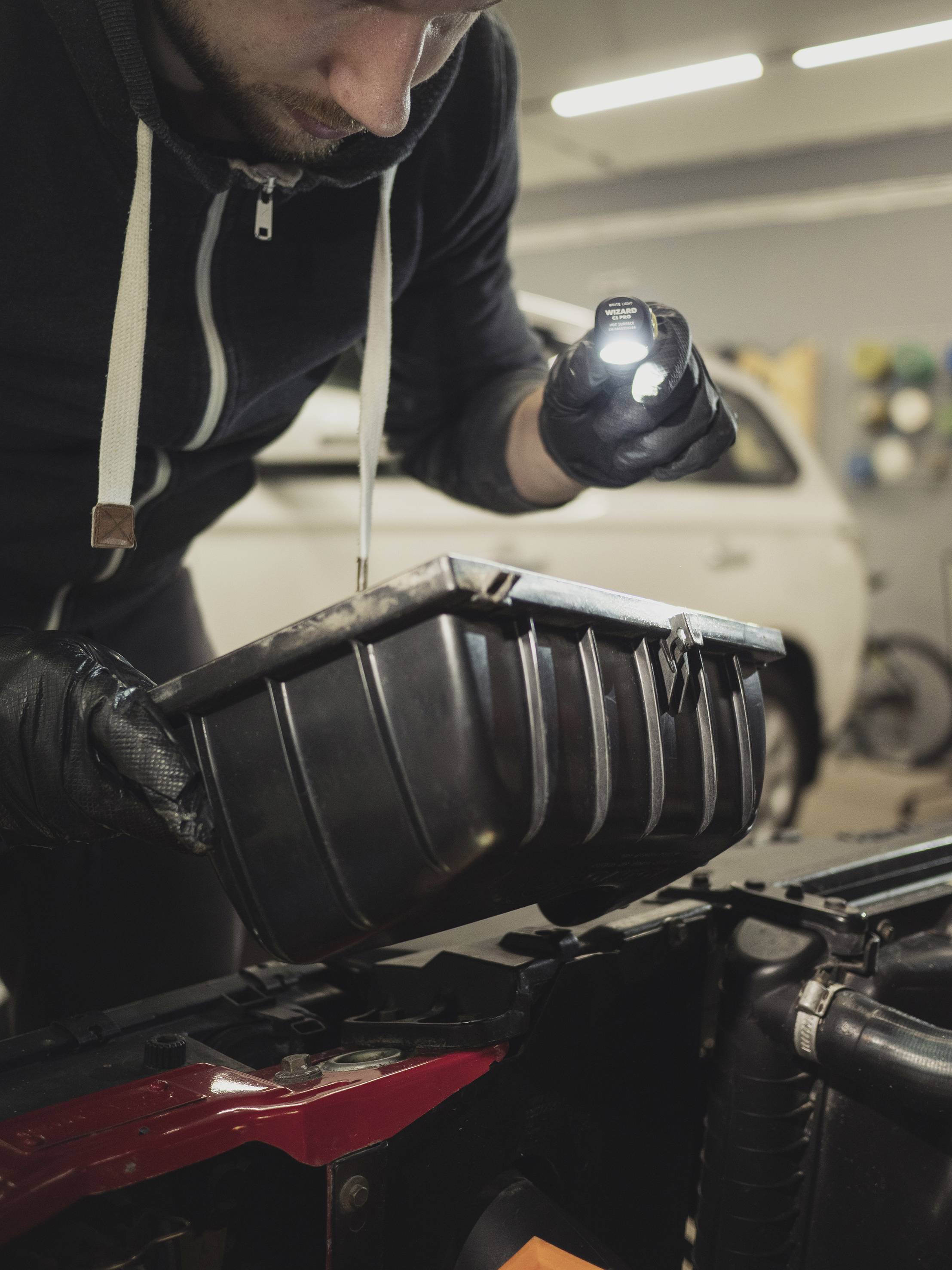 Een persoon inspecteert de motorruimte van een auto met een zaklamp. Ze draagt handschoenen en houdt een open container vast.