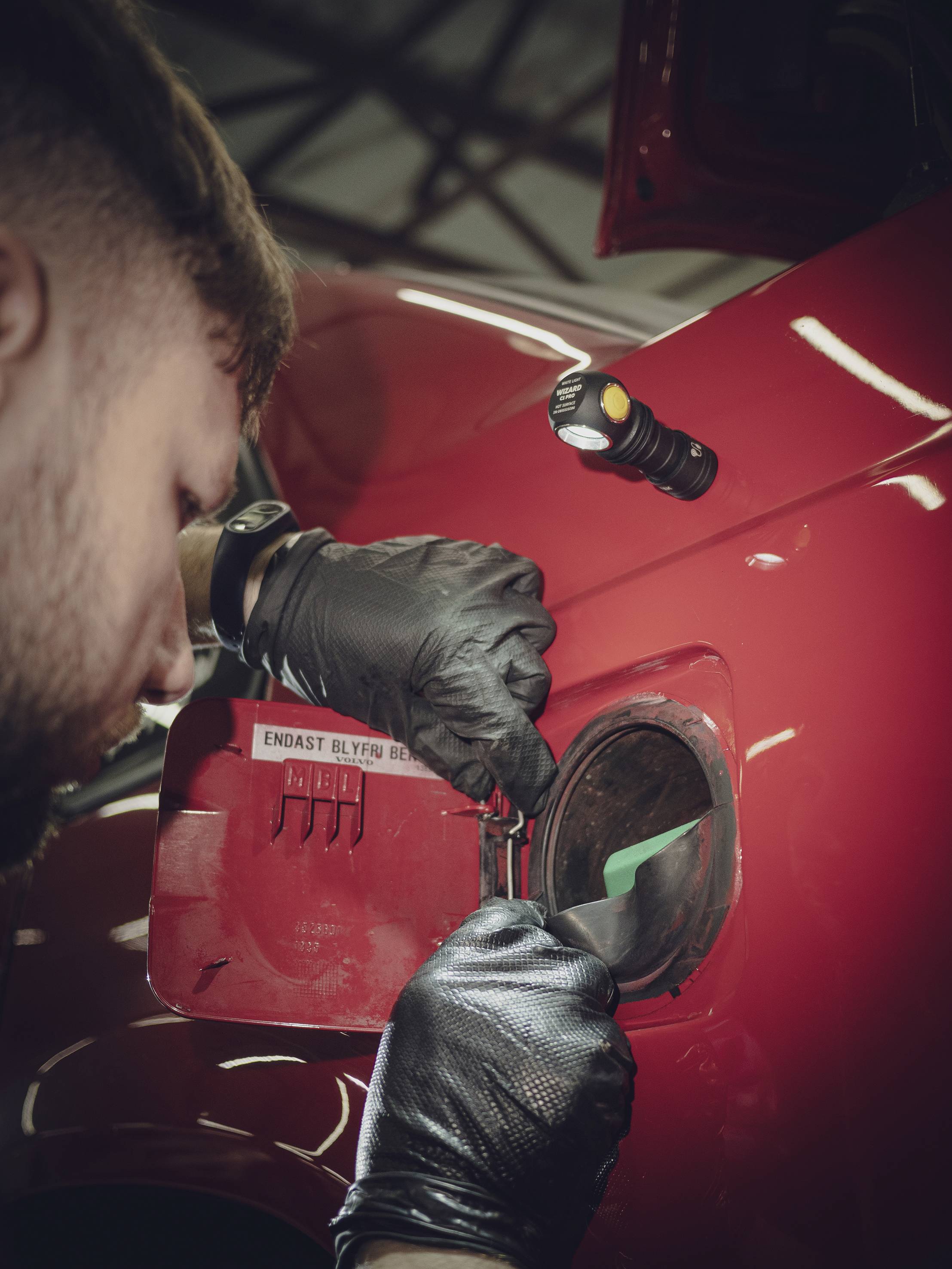 Een man met zwarte handschoenen opent de tankdop van een rode auto in een werkplaatsomgeving.