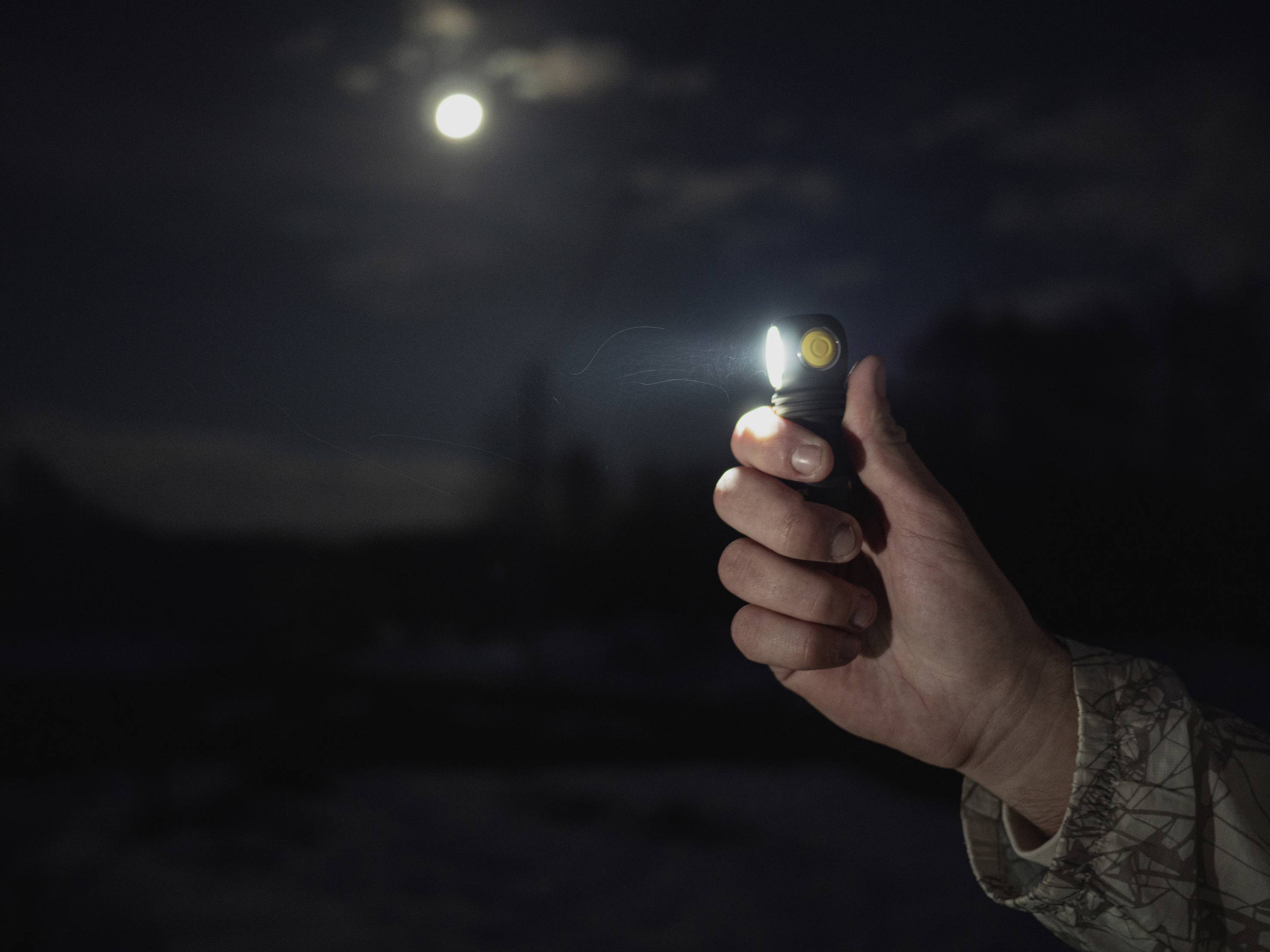 'Een hand houdt een oplichtende zaklamp in het donker, terwijl de volle maan aan de hemel op de achtergrond staat.'