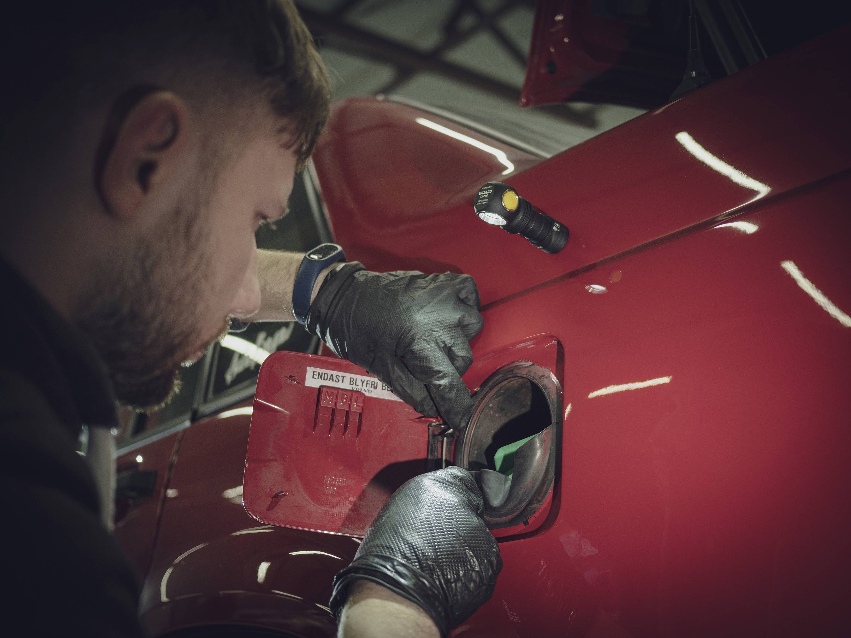 Een man vult een rode auto met brandstof. Hij draagt zwarte handschoenen en gebruikt een zaklamp om de tankopening te verlichten.
