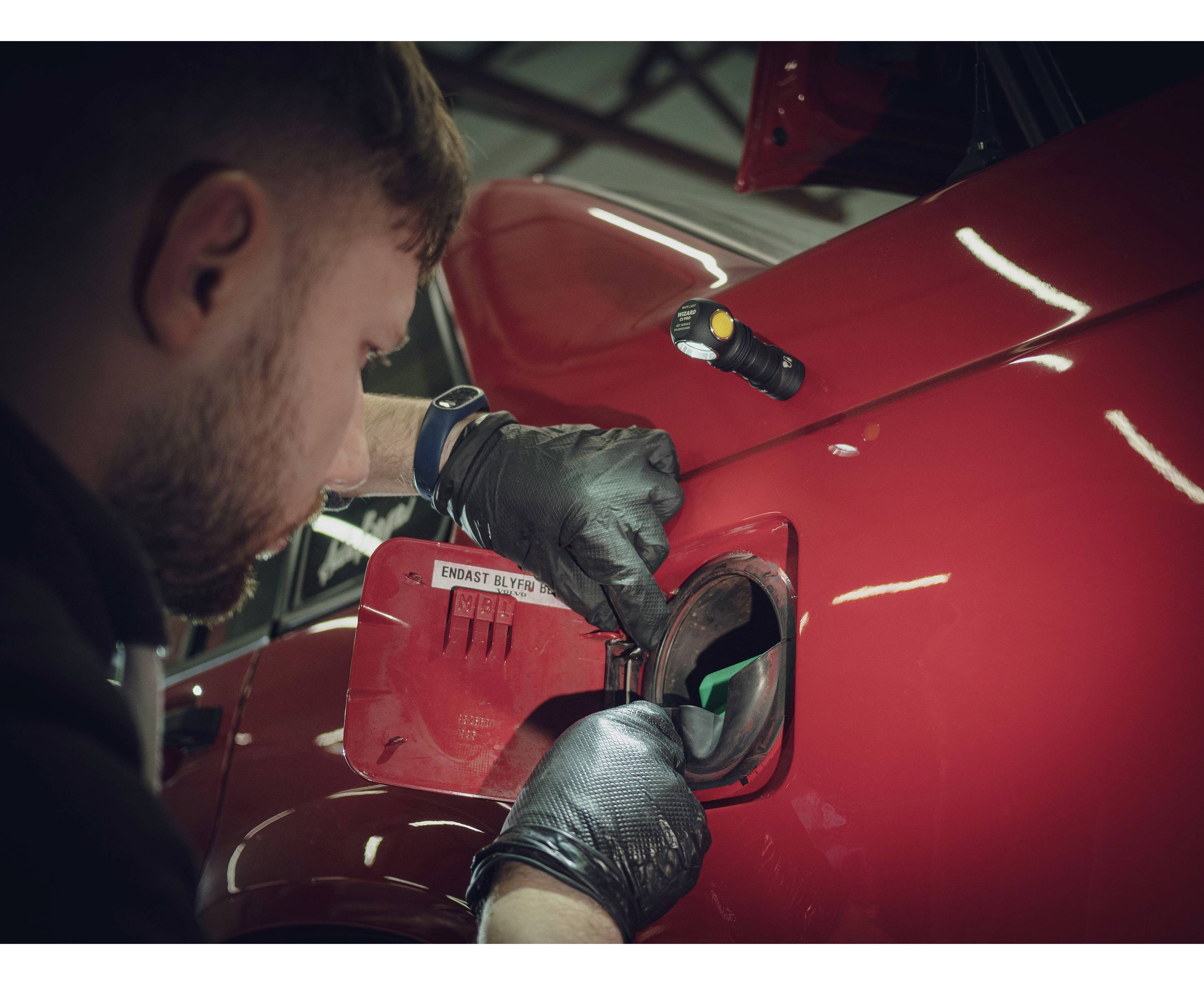 Een monteur met zwarte handschoenen inspecteert de geopende tankdop van een rode auto in een werkplaats.