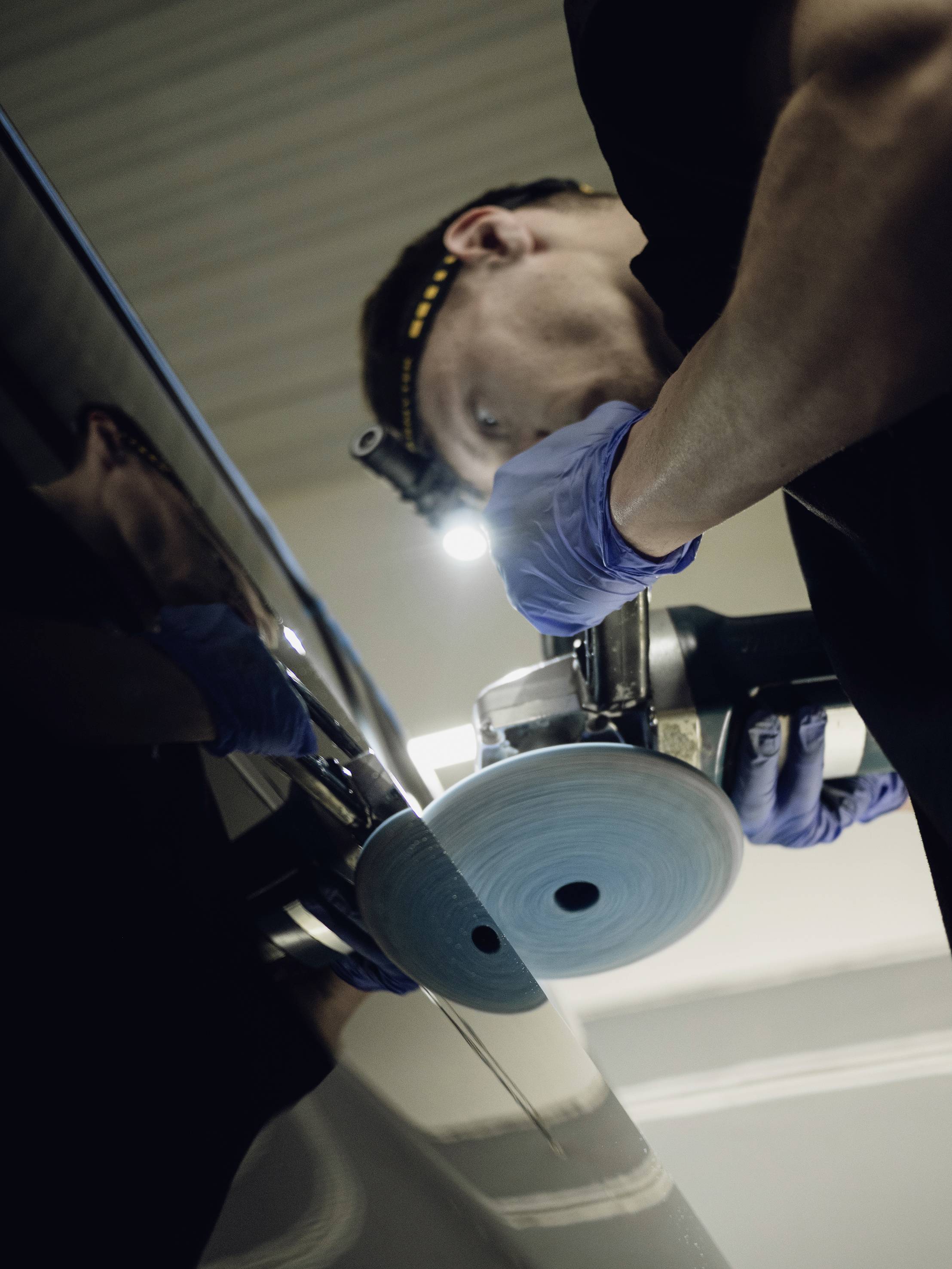 Een monteur polijst de autolak met een polijstmachine. Hij draagt blauwe handschoenen en een hoofdlamp voor beter zicht.