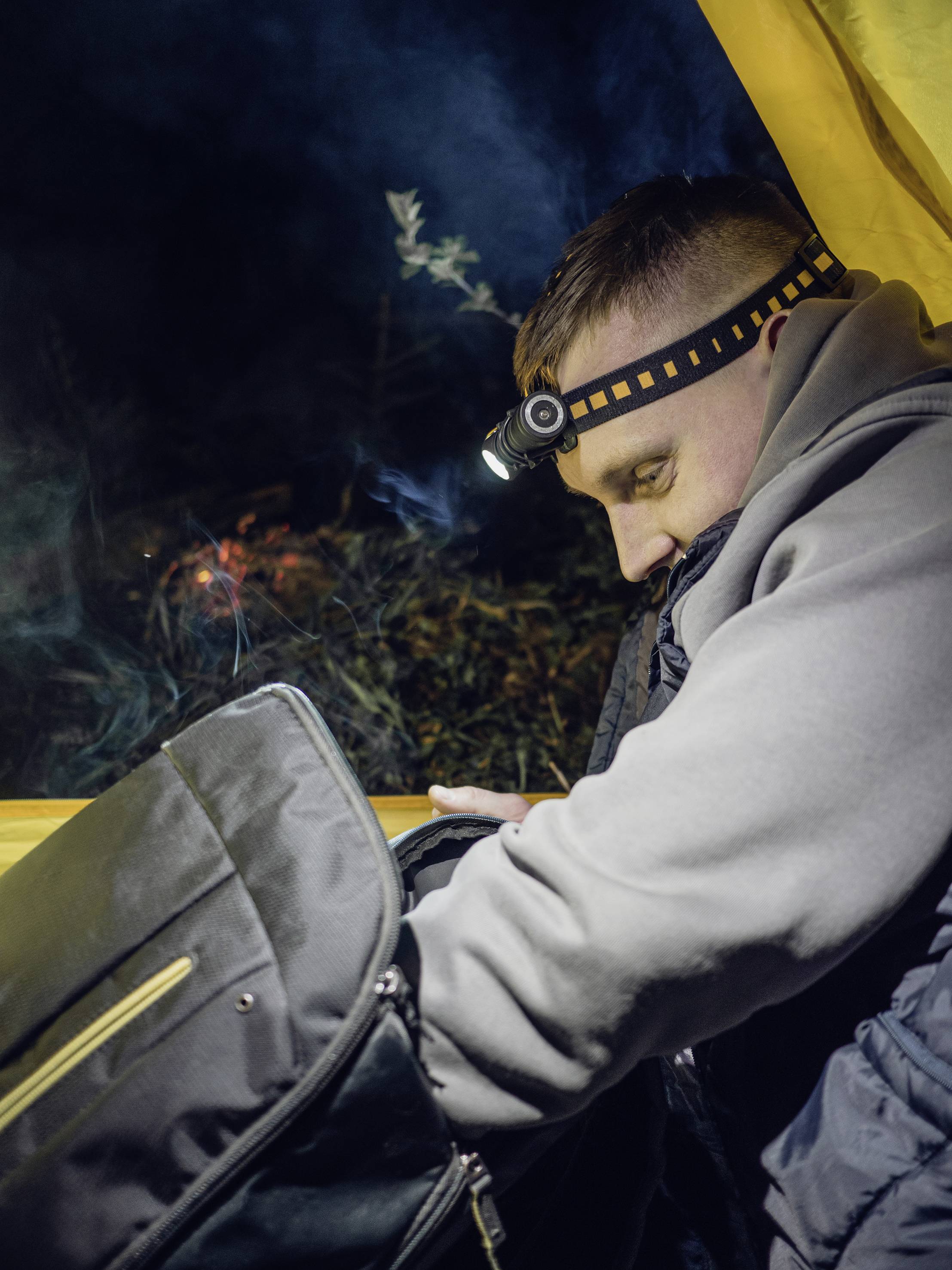 Een man met een hoofdlamp in een tent doorzoekt een tas. Het is nacht, en op de achtergrond zijn bomen en zwak licht zichtbaar.