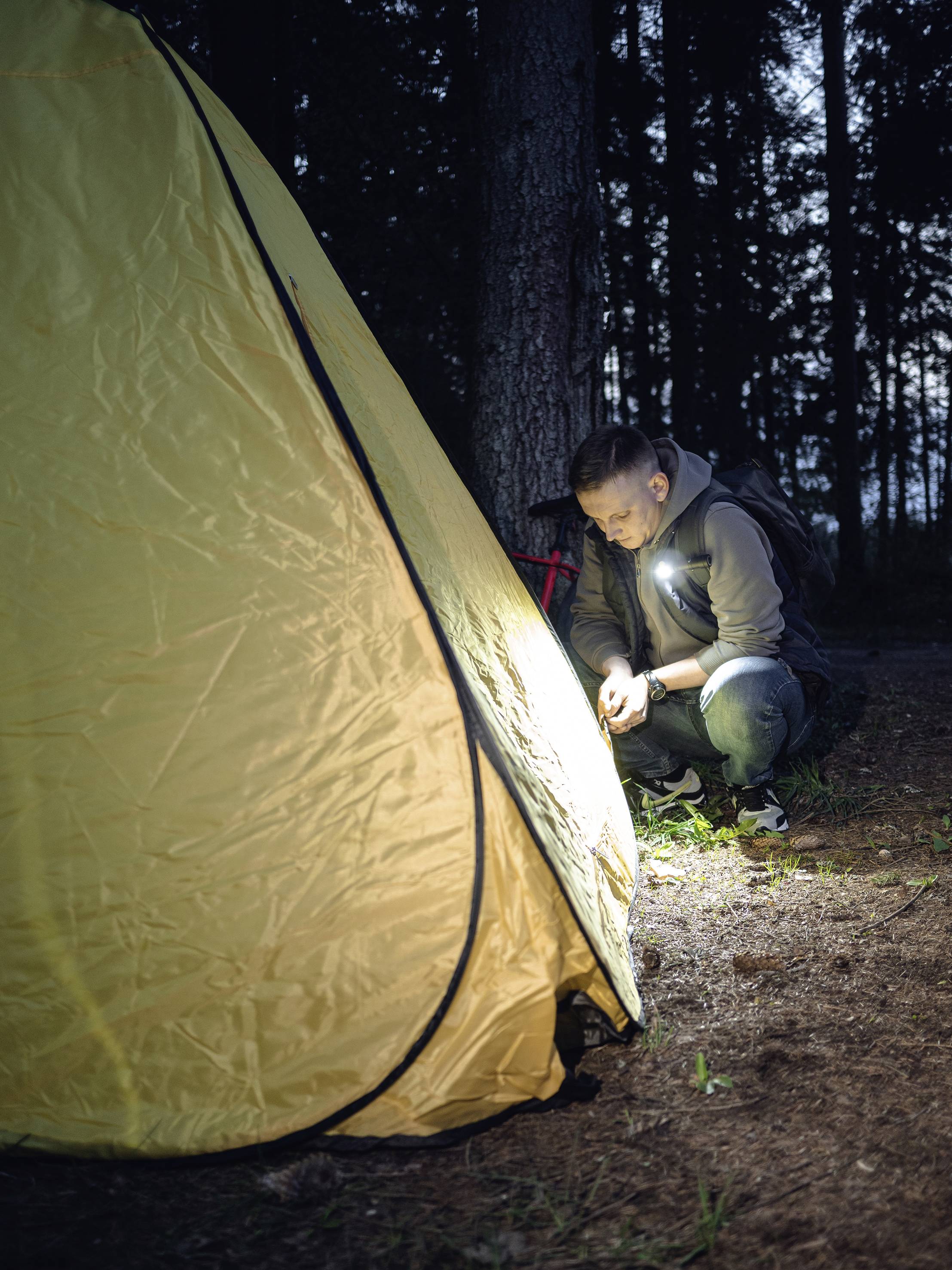 Een persoon knielt naast een geel tent in het bos en controleert iets op de grond met een zaklamp in het donker.