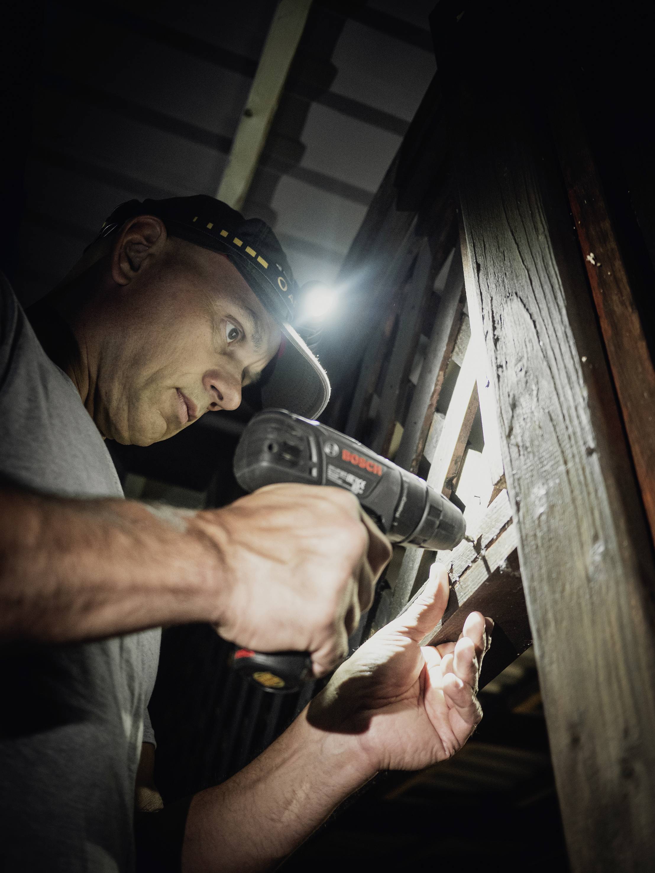 Een man met een hoofdlamp gebruikt een accuschroevendraaier om hout te bewerken. Omgevingslicht is zwak, focus op ambachtelijk werk.