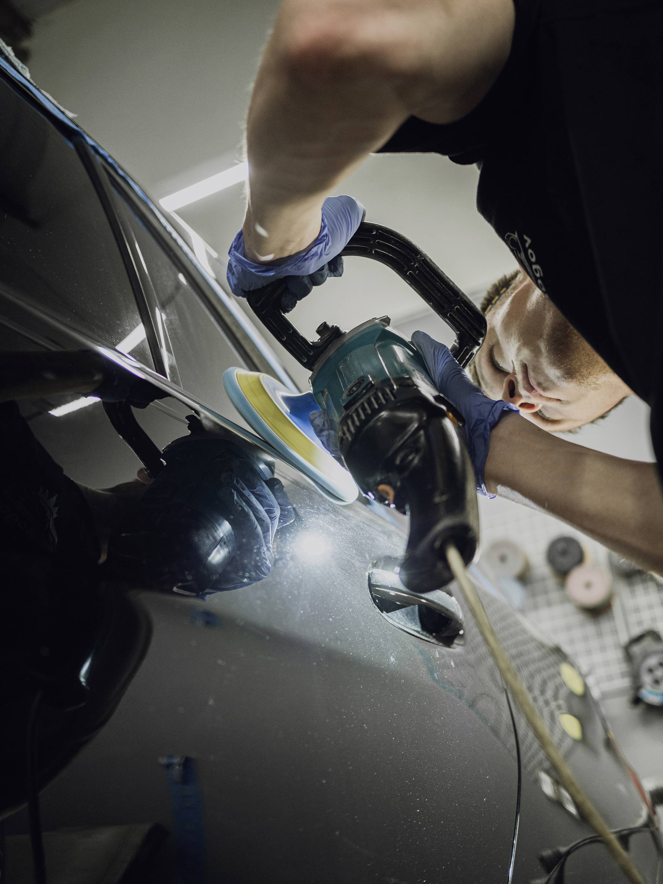 Een persoon polijst een donkere autolak met een machine in een werkplaats, draagt daarbij blauwe handschoenen.