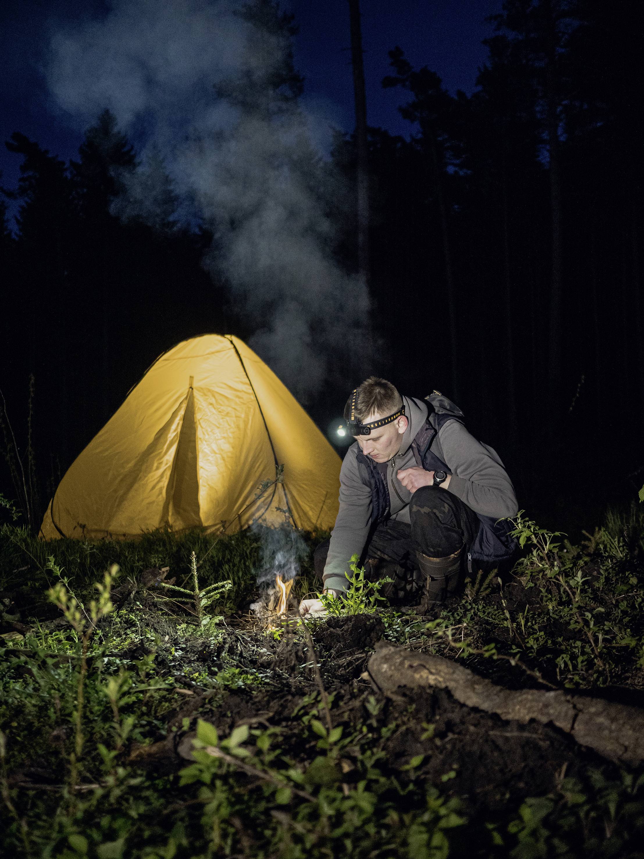 Een man steekt een kampvuur aan voor een geel tent in het donkere bos, draagt een hoofdlamp; nachtopname.