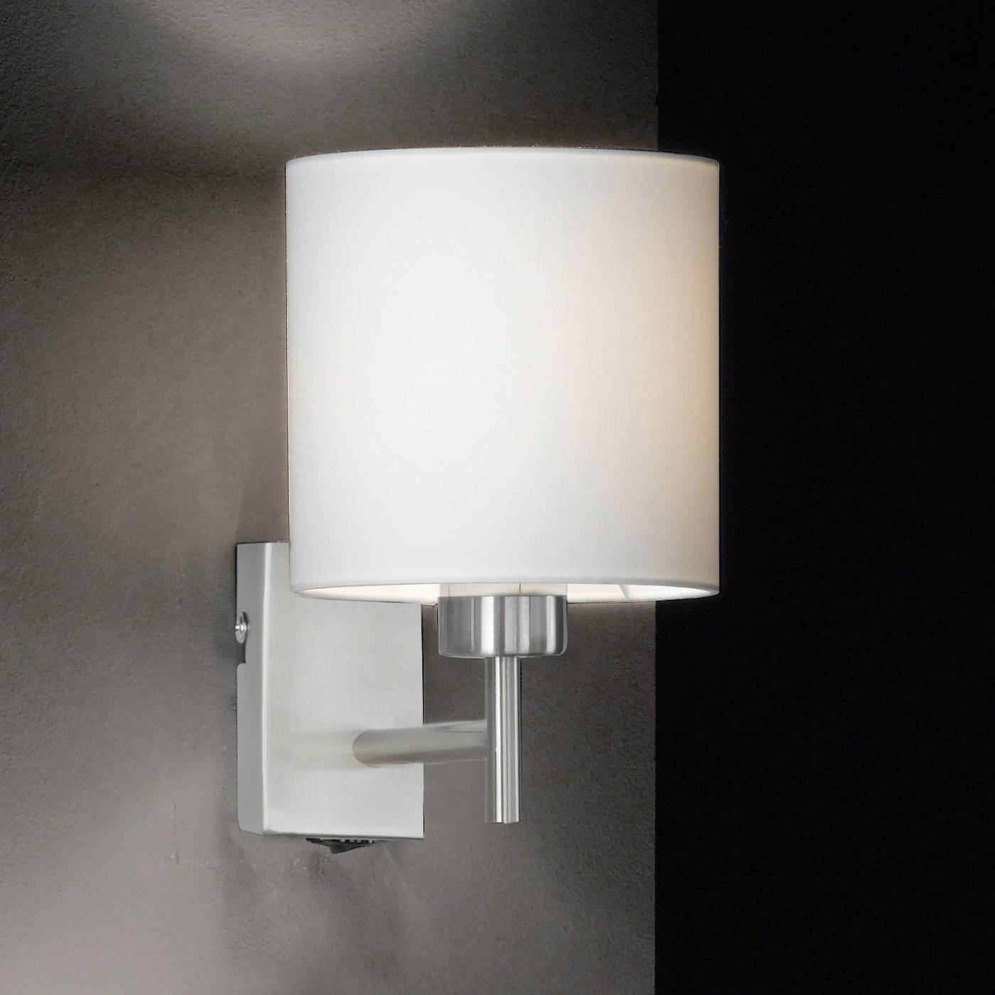 Een moderne wandlamp met een witte, cilindrische lampenkap, gemonteerd aan een metalen bevestiging tegen een donkere muur.