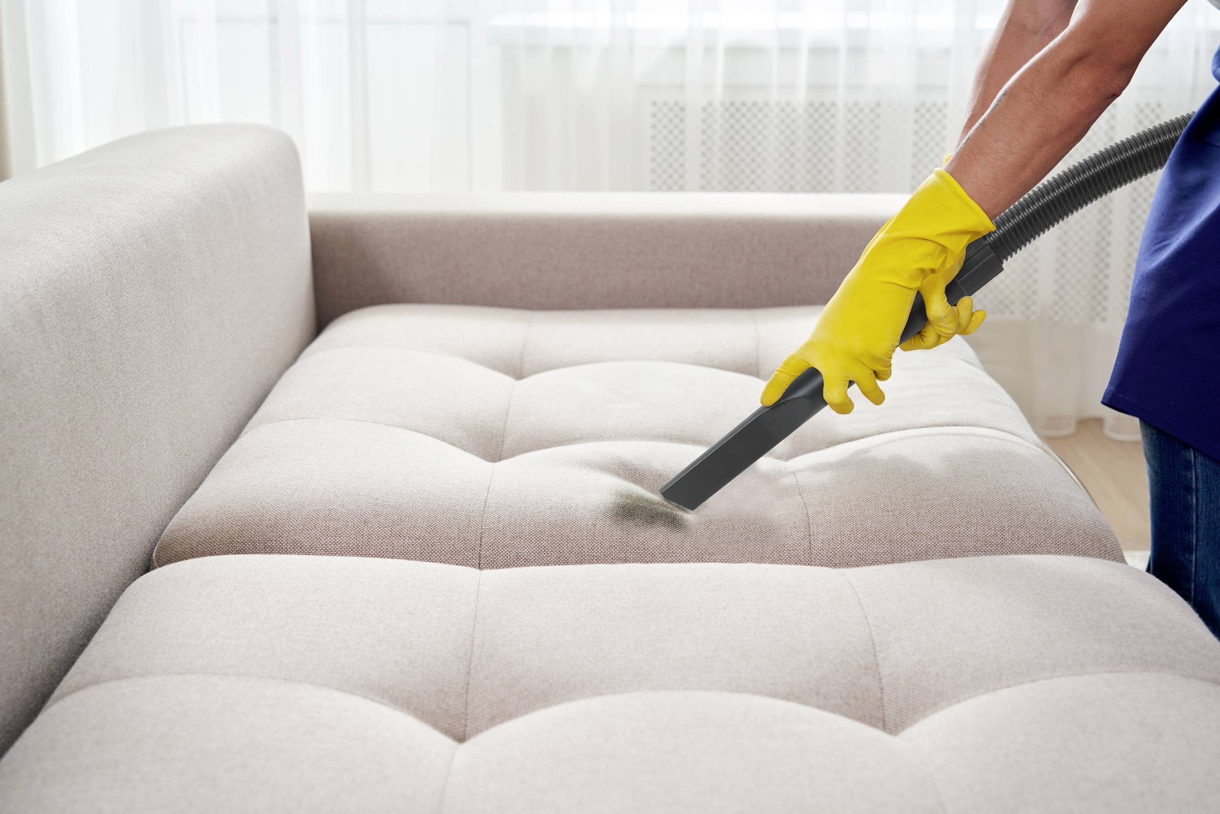 Een persoon reinigt een beige bank met een stofzuiger. Ze draagt gele schoonmaakhandschoenen.