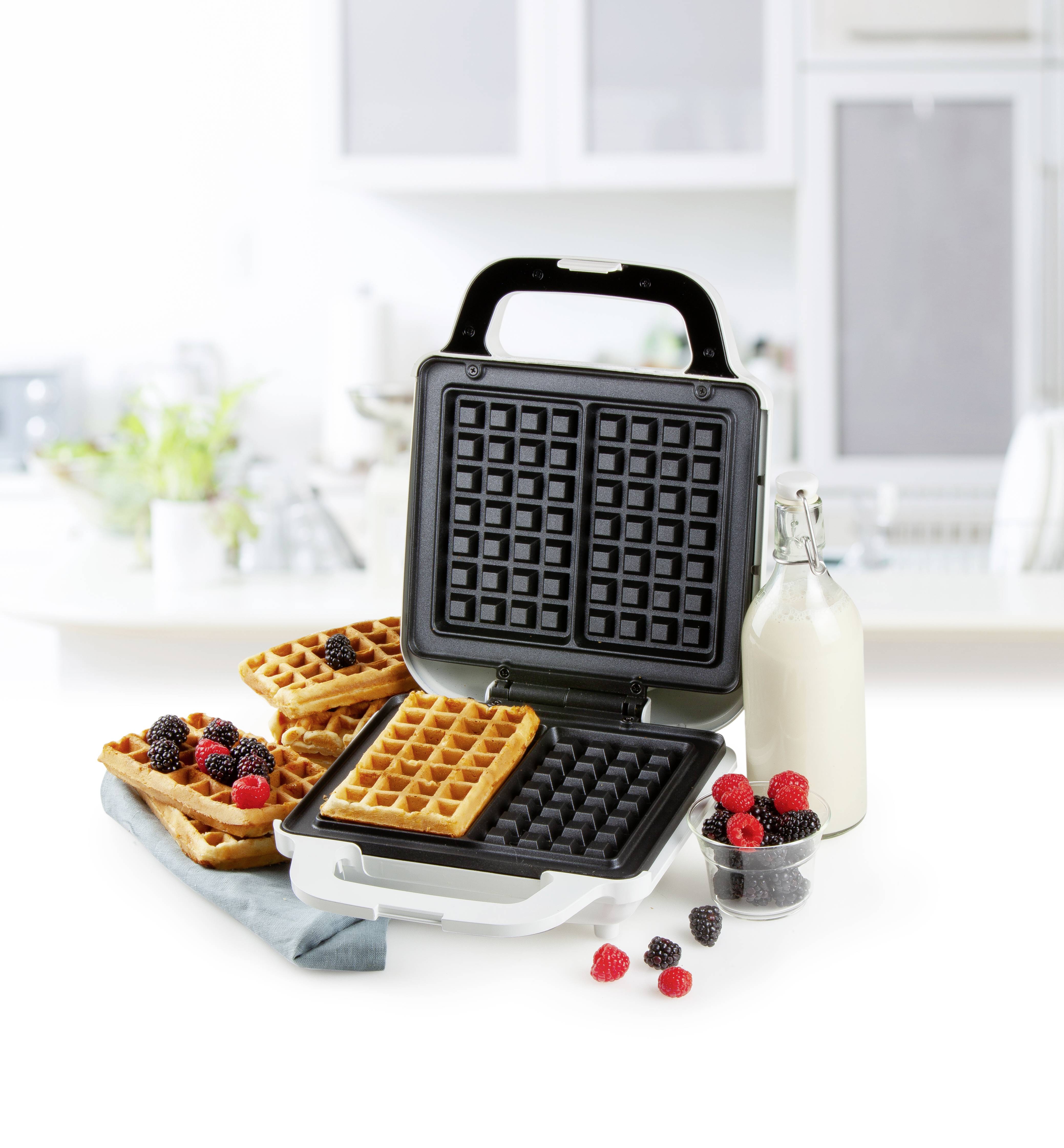 Een wafelijzer met geopend deksel, met daarin twee goudbruine wafels. Ernaast verse bessen en een fles melk in een moderne keuken.
