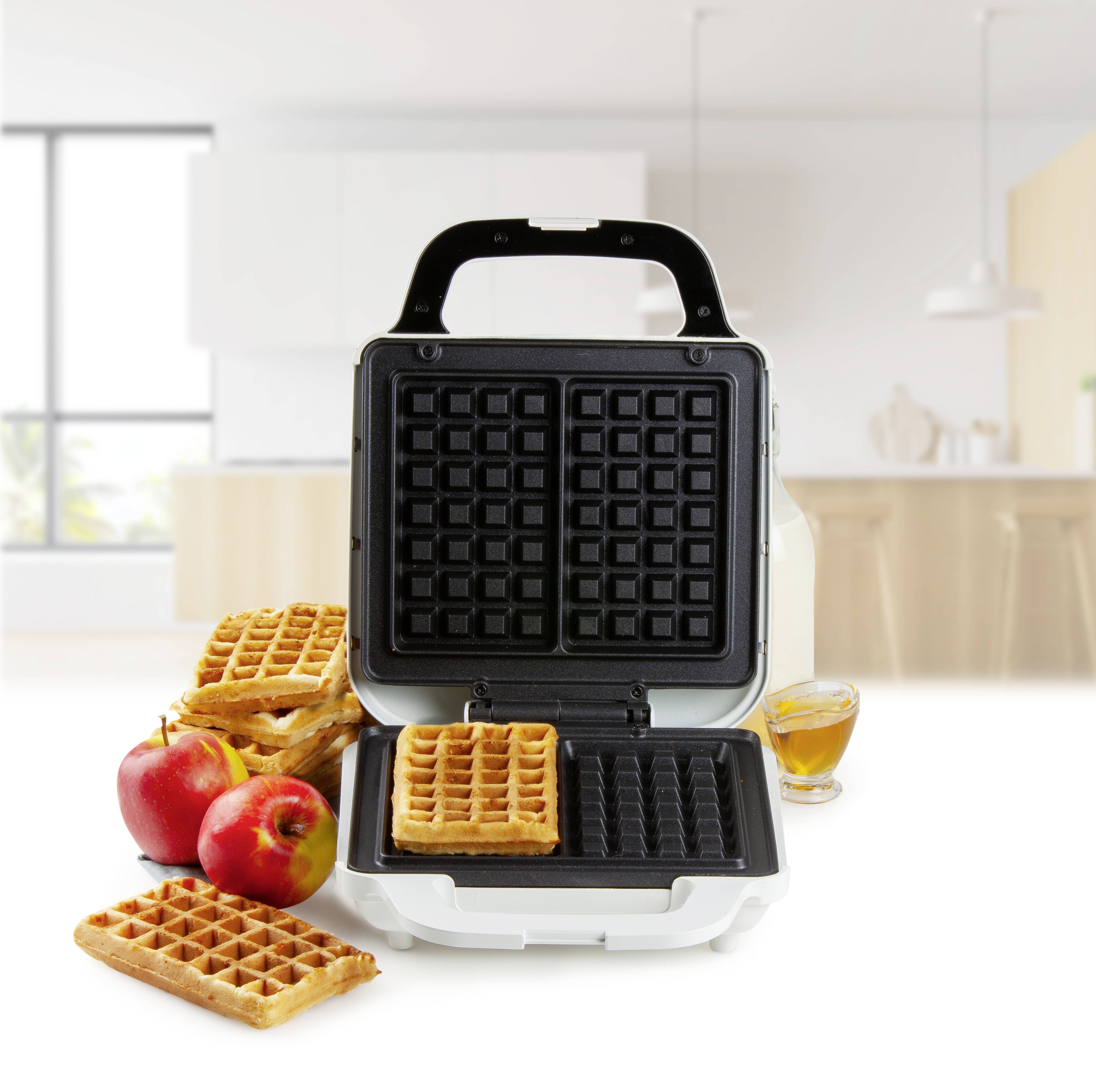 Een wafelijzer in een keuken, omringd door wafels, appels en een glas siroop.