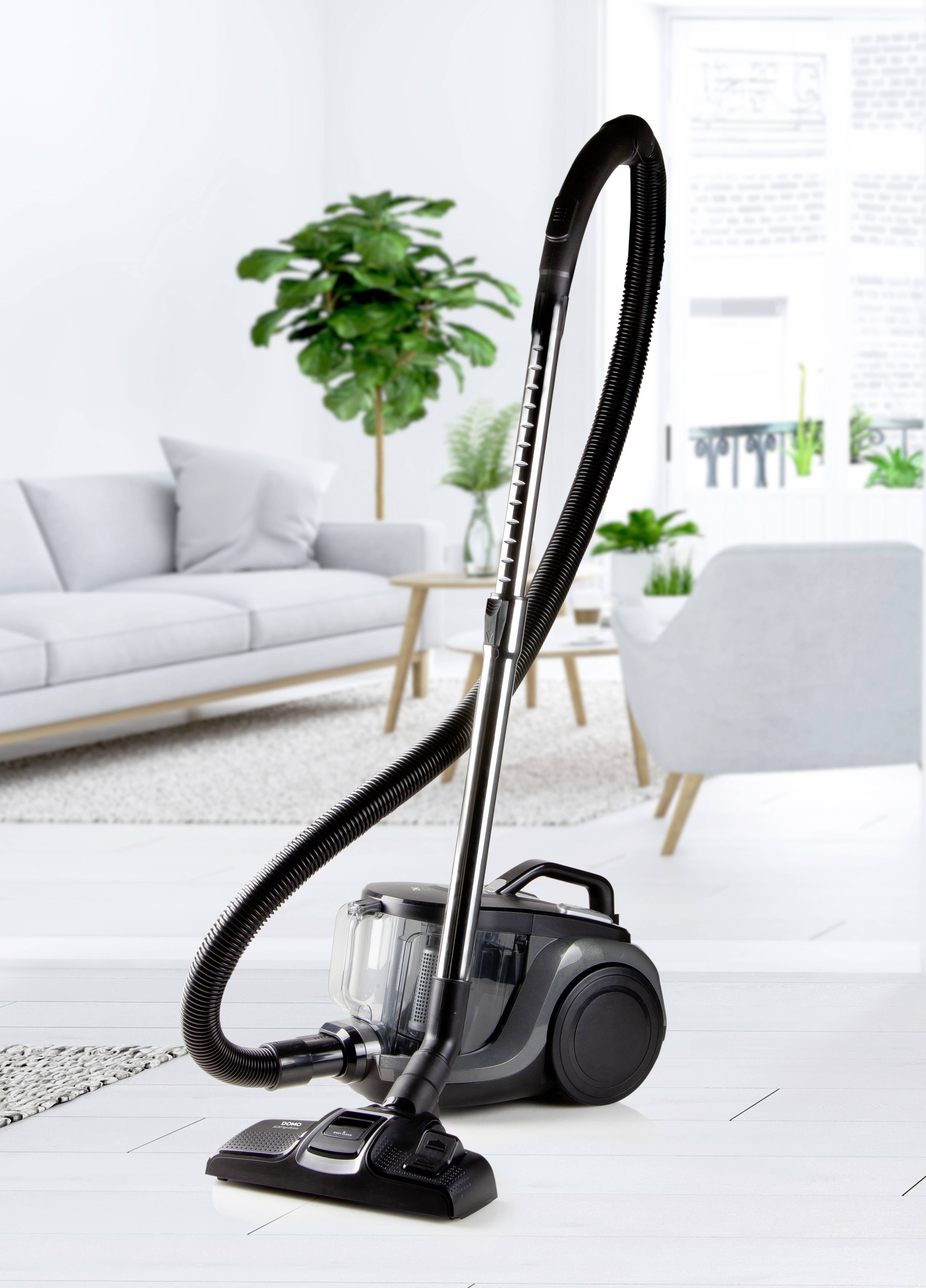 Stofzuiger staat in de woonkamer op een witte vloer, dicht bij een grijze bank en een groene plant, daglicht van grote ramen.
