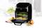 Een zwarte airfryer met geopende deur toont voedsel op twee roosterlagen, omringd door gebak en appels in een keuken.