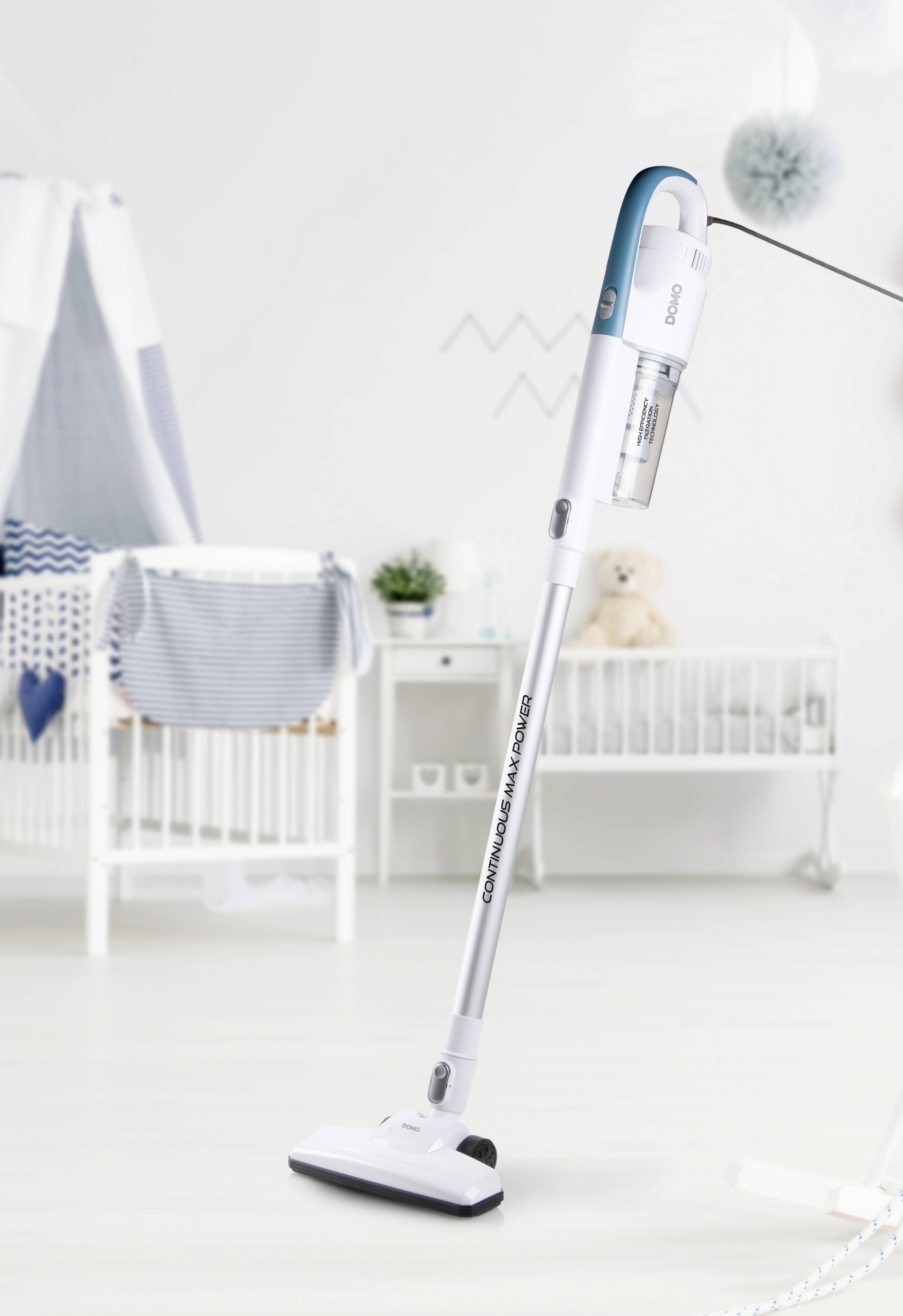 Een draadloze stofzuiger staat rechtop op een lichte vloer in een kinderkamer met een wiegend wiegje en subtiele decoraties.