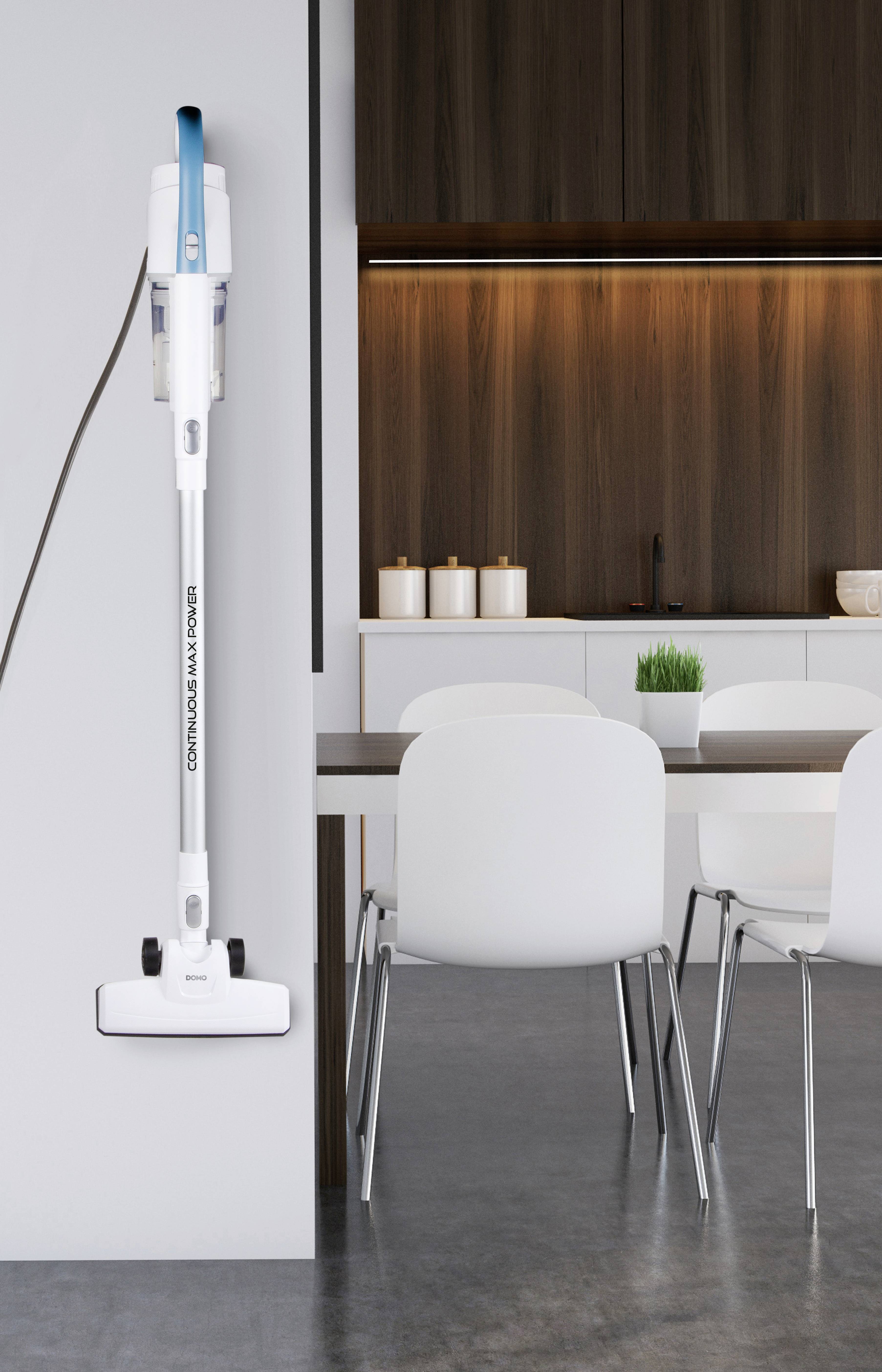 Een witte stofzuiger hangt aan een lichte muur in een moderne keuken met houten keukenkastjes, witte stoelen en een tafel.