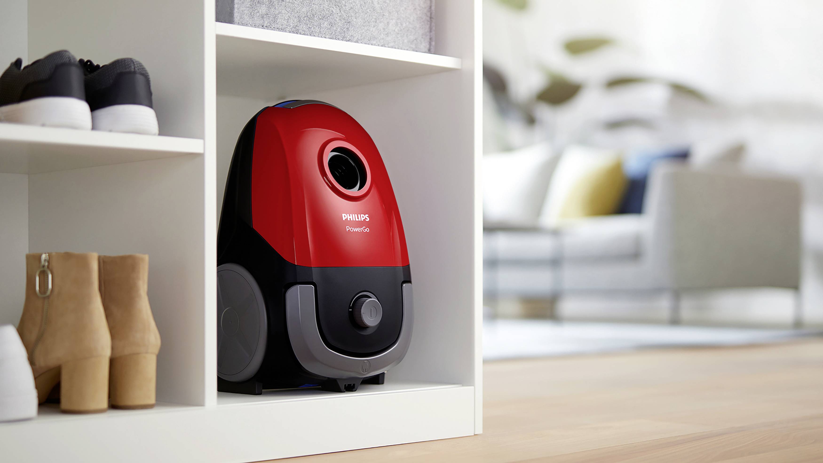 Stofzuiger in een rek, naast schoenen, in de woonkamer. De stofzuiger is rood en zwart. De ruimte toont een moderne inrichting.