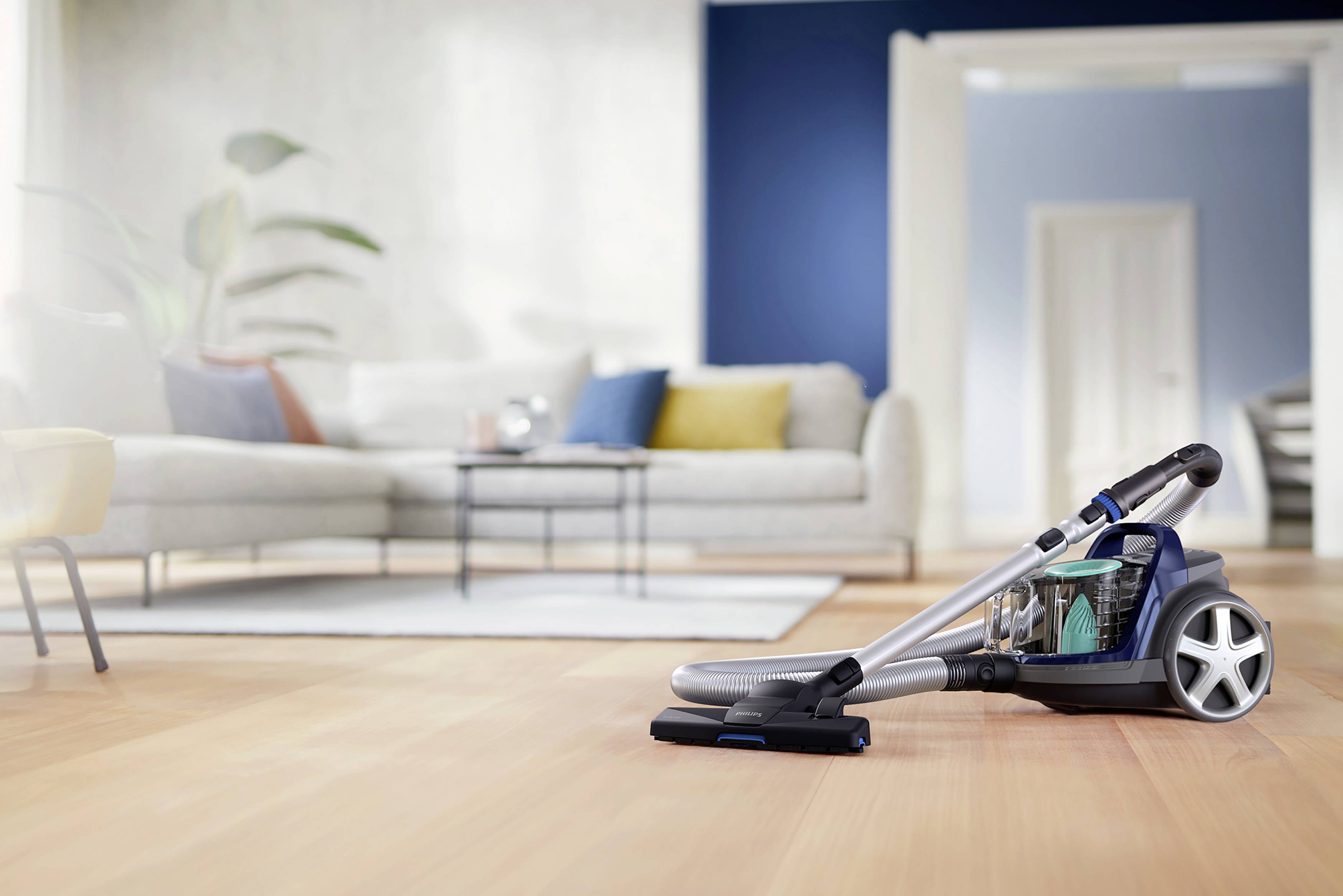 Een stofzuiger op een houten vloer in een modern ingericht woonkamer met bank, kussens en een klein tafeltje op de achtergrond.