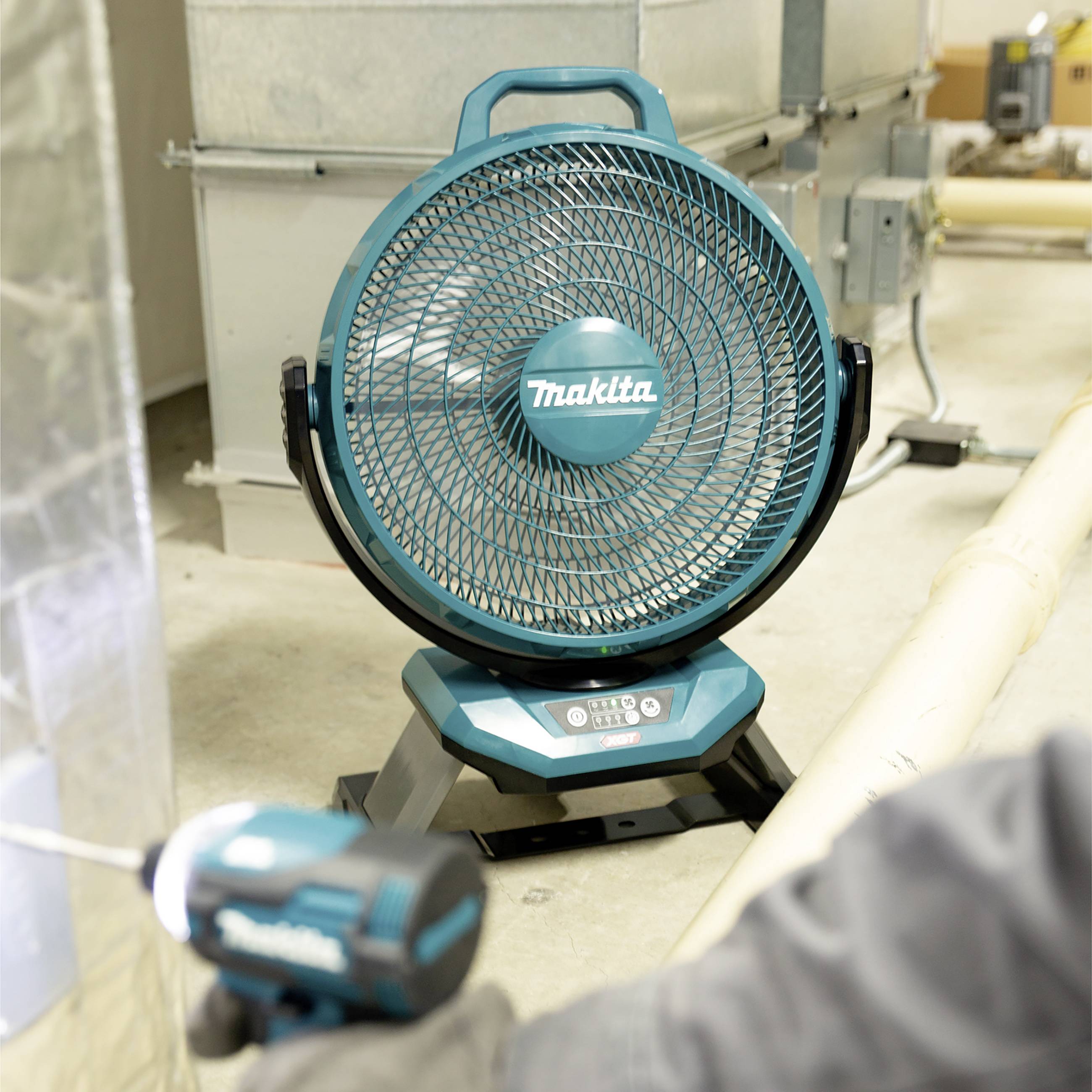 Een blauwe, draagbare ventilator staat in een industriële ruimte. Op de voorgrond houdt een persoon een gereedschap vast.