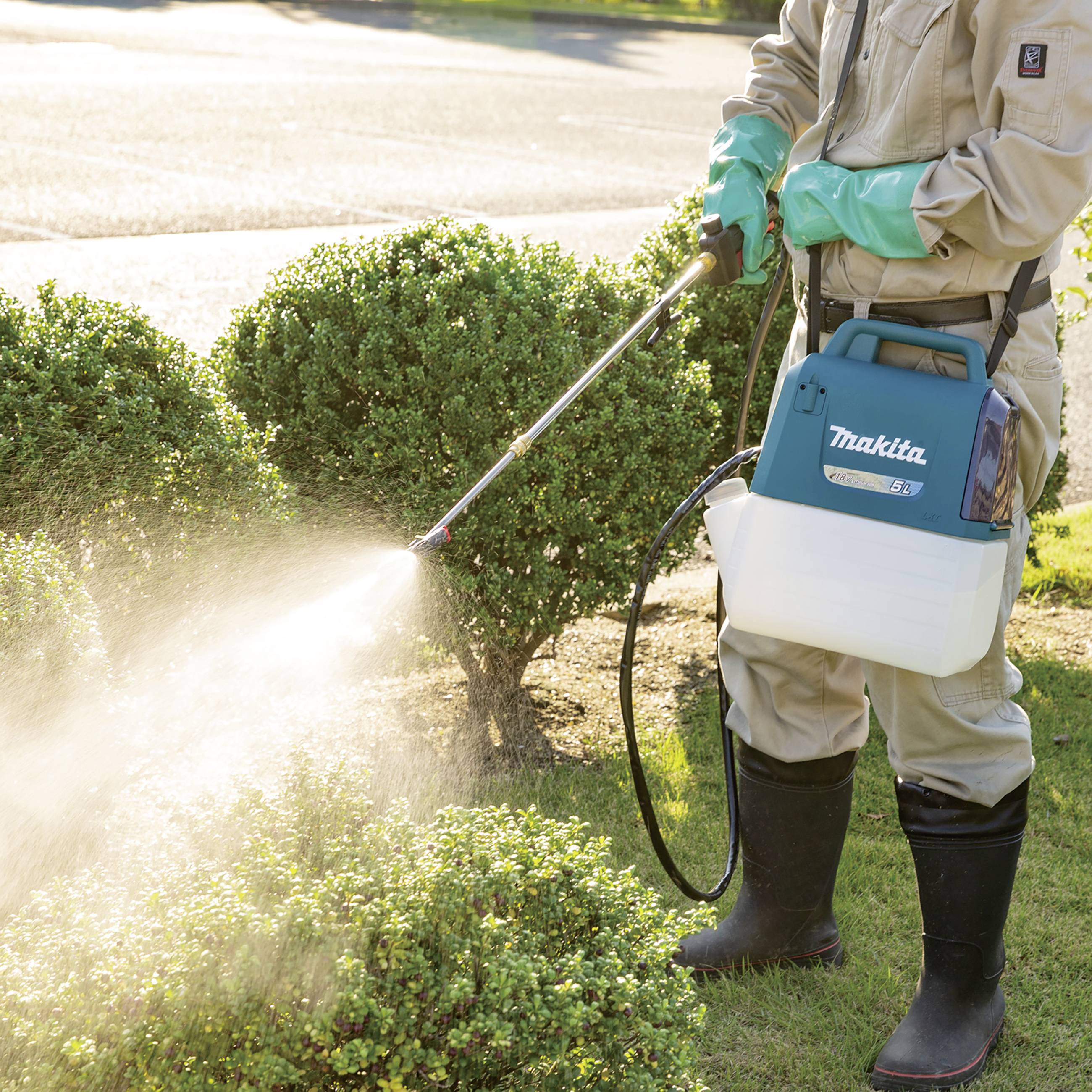 Een persoon besproeit struiken met een spuitapparaat in de tuin. Ze draagt handschoenen en rubberlaarzen. Zonlicht schijnt op de planten.