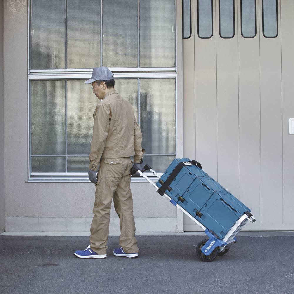 Een persoon in werkkleding trekt een blauwe transportkar met gereedschapskisten voor een gebouw.